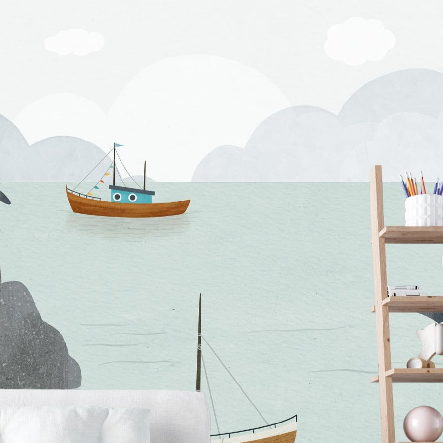 wall adorned with the Kids Nautical Mural Wallpaper, displaying the artful depiction of a coastal landscape. The mural creates a serene ambiance with its pastel sky, calm sea, and floating boats, all watched over by seagulls in flight and perched atop the rocks, making it a picturesque backdrop for a child's play area or bedroom.