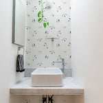 A small bathroom decorated with the Birds By the Sea Wallpaper, featuring a pattern of seagulls in flight on a light background. The bathroom includes a modern white sink on a marble countertop, with a hanging plant adding a touch of greenery under a skylight.
