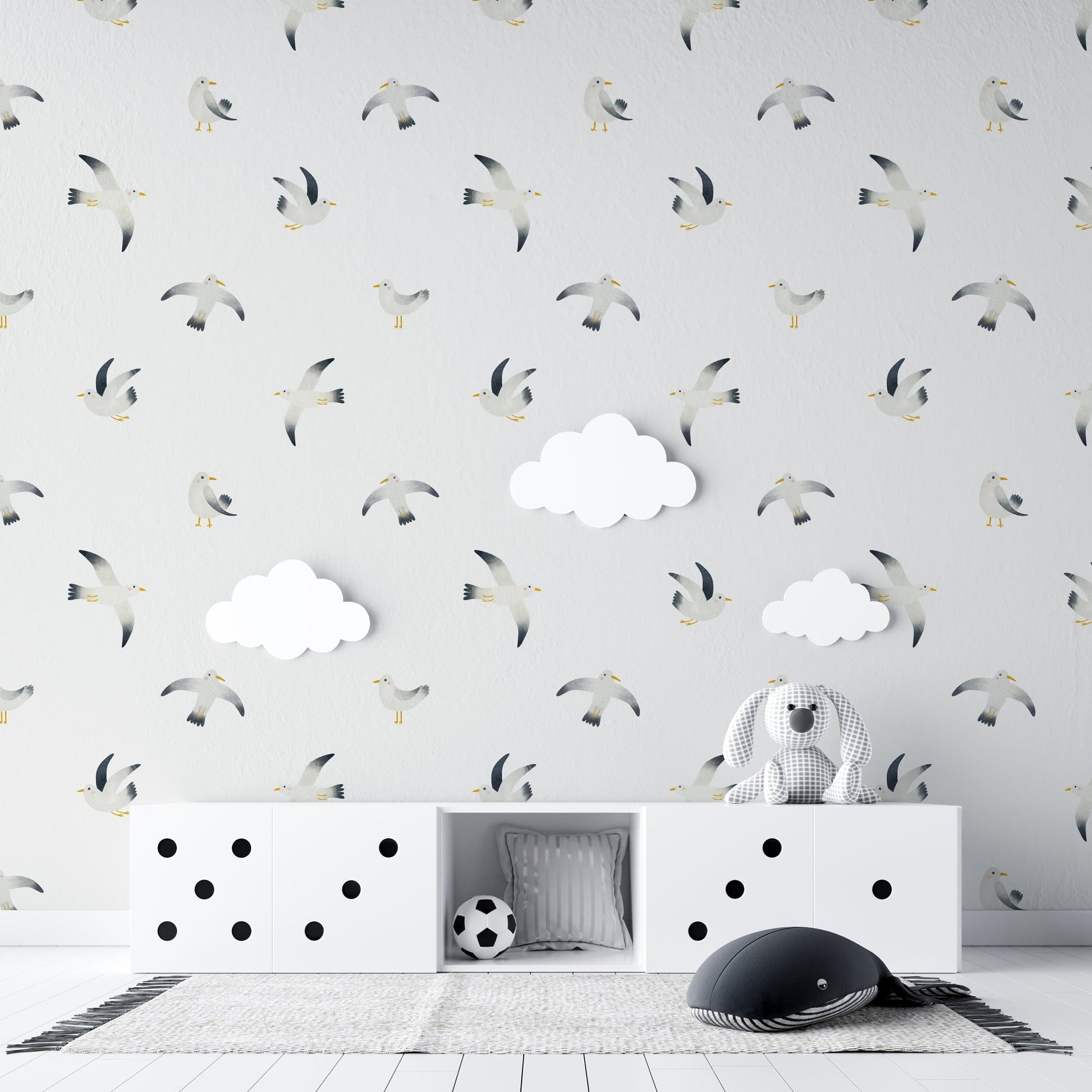 A playful setting in a child’s room featuring the Birds By the Sea Wallpaper. The room is furnished with a white storage unit adorned with black polka dots and fluffy white clouds hanging on the walls, complementing the flying seagull pattern.