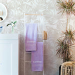 A vertical image showcasing a corner of an interior space with a distinct white wallpaper adorned with large, embossed monstera leaf patterns. In the foreground, a towel rack with branded purple towels is visible alongside a plant and a wicker basket.