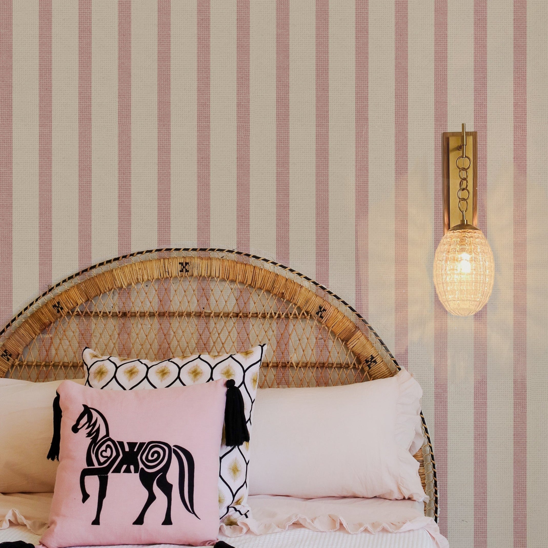 A serene bedroom setting featuring Pink Textured Striped Wallpaper with alternating light and dark pink stripes on a textured background. The room is styled with a rattan headboard, white bedding, and a decorative pink cushion with a horse motif, complemented by a modern wall-mounted light.