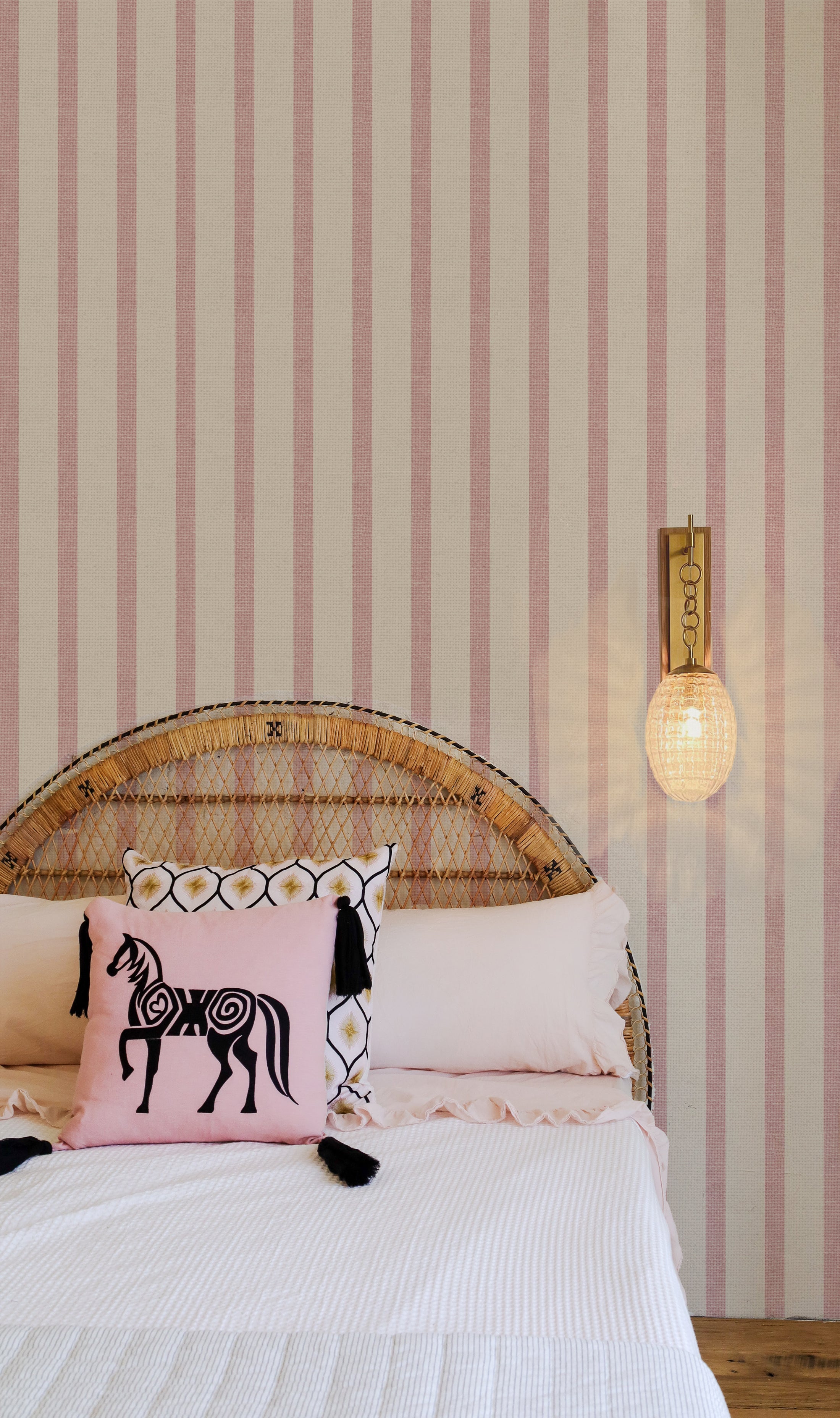 A serene bedroom setting featuring Pink Textured Striped Wallpaper with alternating light and dark pink stripes on a textured background. The room is styled with a rattan headboard, white bedding, and a decorative pink cushion with a horse motif, complemented by a modern wall-mounted light.