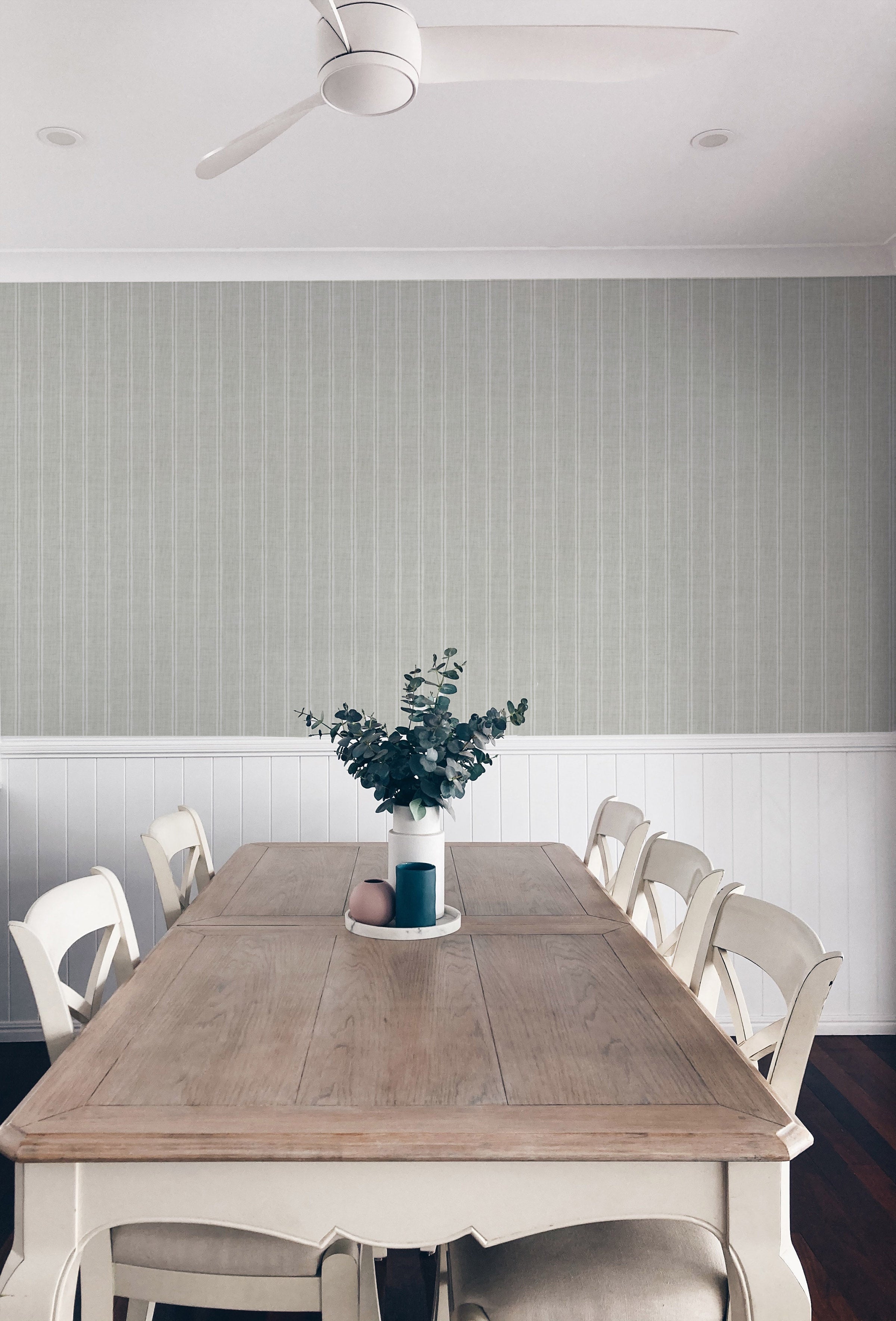 A rustic dining room scene is brought to life with the Olive Textured Striped Wallpaper, providing a serene backdrop with its subtle, textured vertical stripes in muted olive tones. The wallpaper pairs beautifully with the classic wooden dining table and white chairs, under a modern white ceiling fan.
