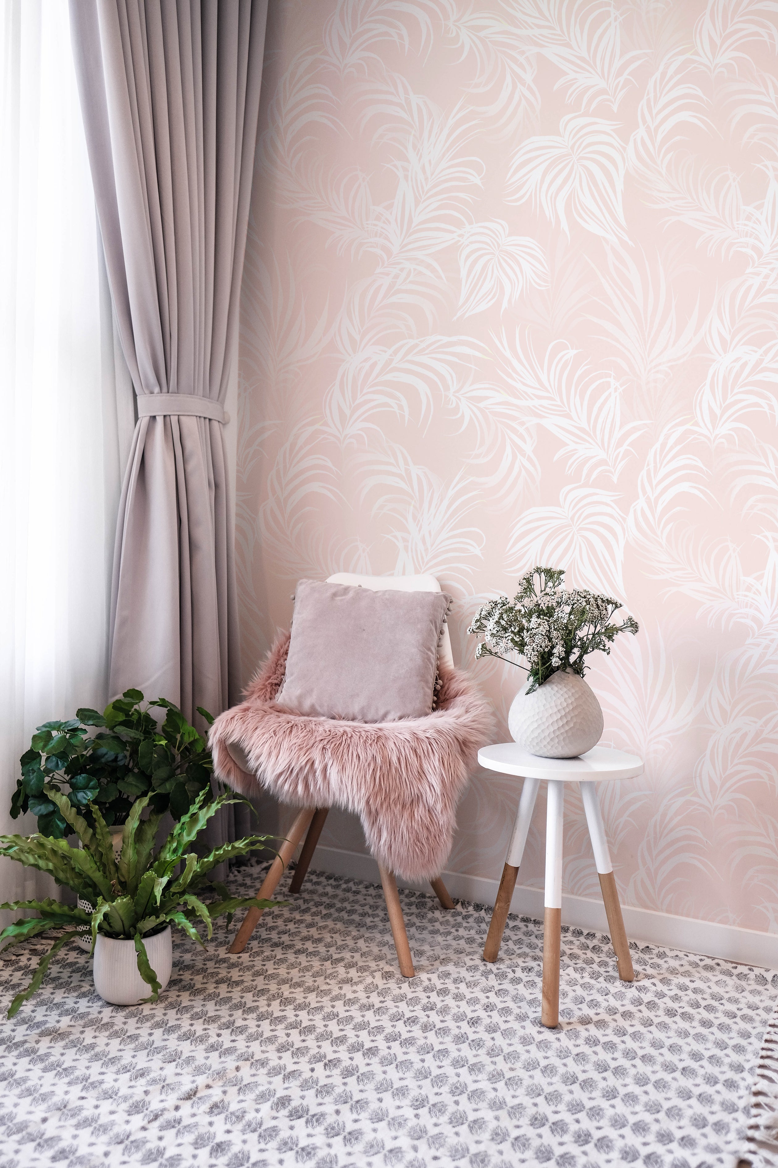 A cozy nook enhanced by the "Tropical Champagne Wallpaper," displaying a soothing palette of blush pink with white tropical foliage designs. This wallpaper brings a touch of understated elegance to the space, beautifully paired with a plush chair, a simple white side table, and soft decorative accents.