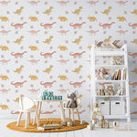Nursery room featuring 'Dino World Kids Wallpaper' with a pattern of pale pink and yellow dinosaurs and delicate foliage on a white background. The room includes a small children's table and chair set, a white shelf with books and toys, enhancing a cozy and imaginative play space.
