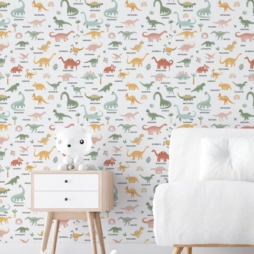 A child's room displaying the Dino World Kids Wallpaper II, which showcases a delightful array of labeled dinosaurs in muted pastels on a white backdrop adorned with green plant motifs. The setting includes a modern white chair and a wooden toy cabinet.