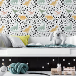 Children's room featuring a wall covered in 'Dino World Kids Wallpaper III' with pastel-colored dinosaur illustrations and black speckles on a white background. The room includes a grey bed with pillows, a plush grey dog toy, and various children's toys on the floor.
