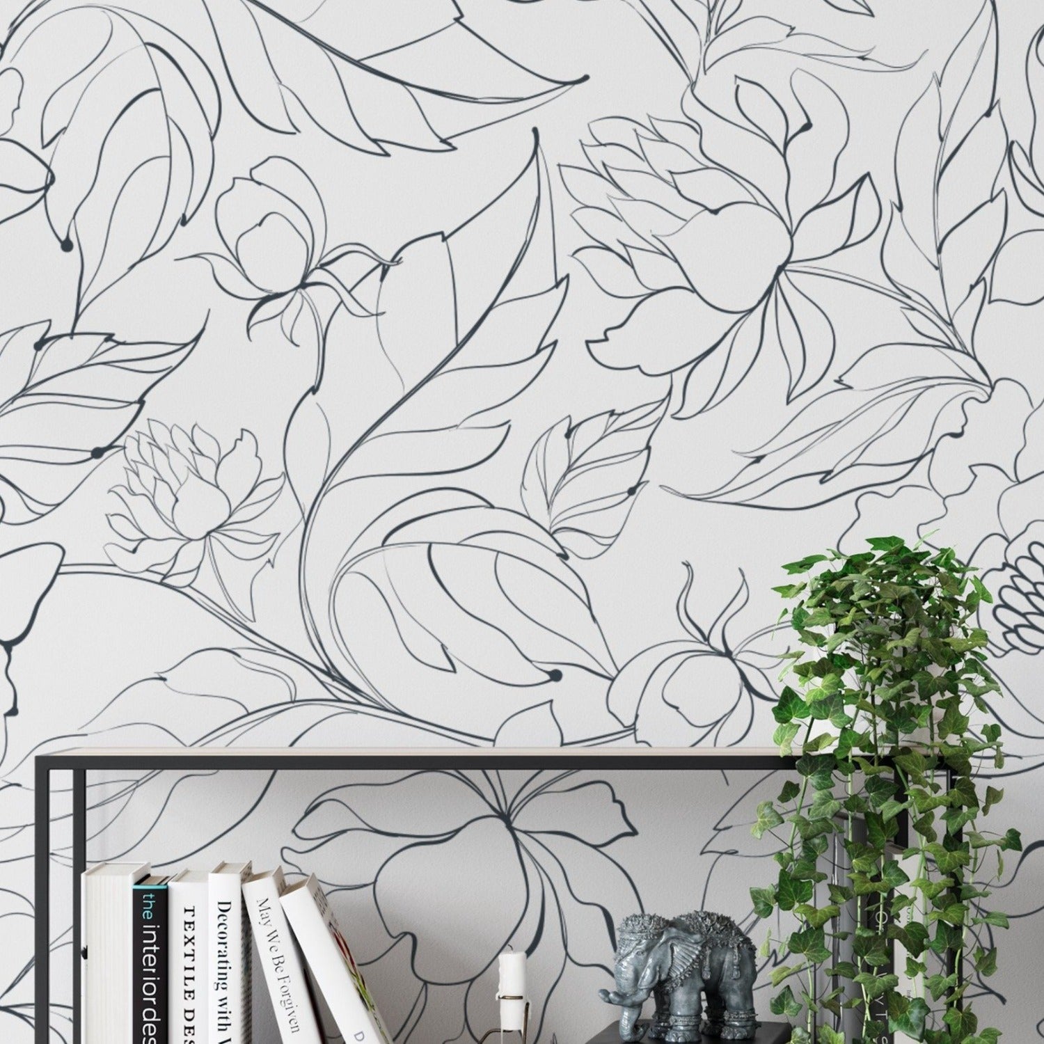 A section of wall with modern black floral wallpaper showcasing bold outlines of various flowers and leaves in a repeating pattern, with a sleek black metal shelf carrying books and a small elephant figurine, beside a climbing ivy plant.