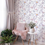 A cozy corner decorated with Watercolor Garden Wallpaper featuring soft purple and pink floral designs on a white background. The area is styled with a plush chair covered in a pink furry throw, a small round table with a vase of flowers, and lush potted plants, creating a tranquil and stylish nook