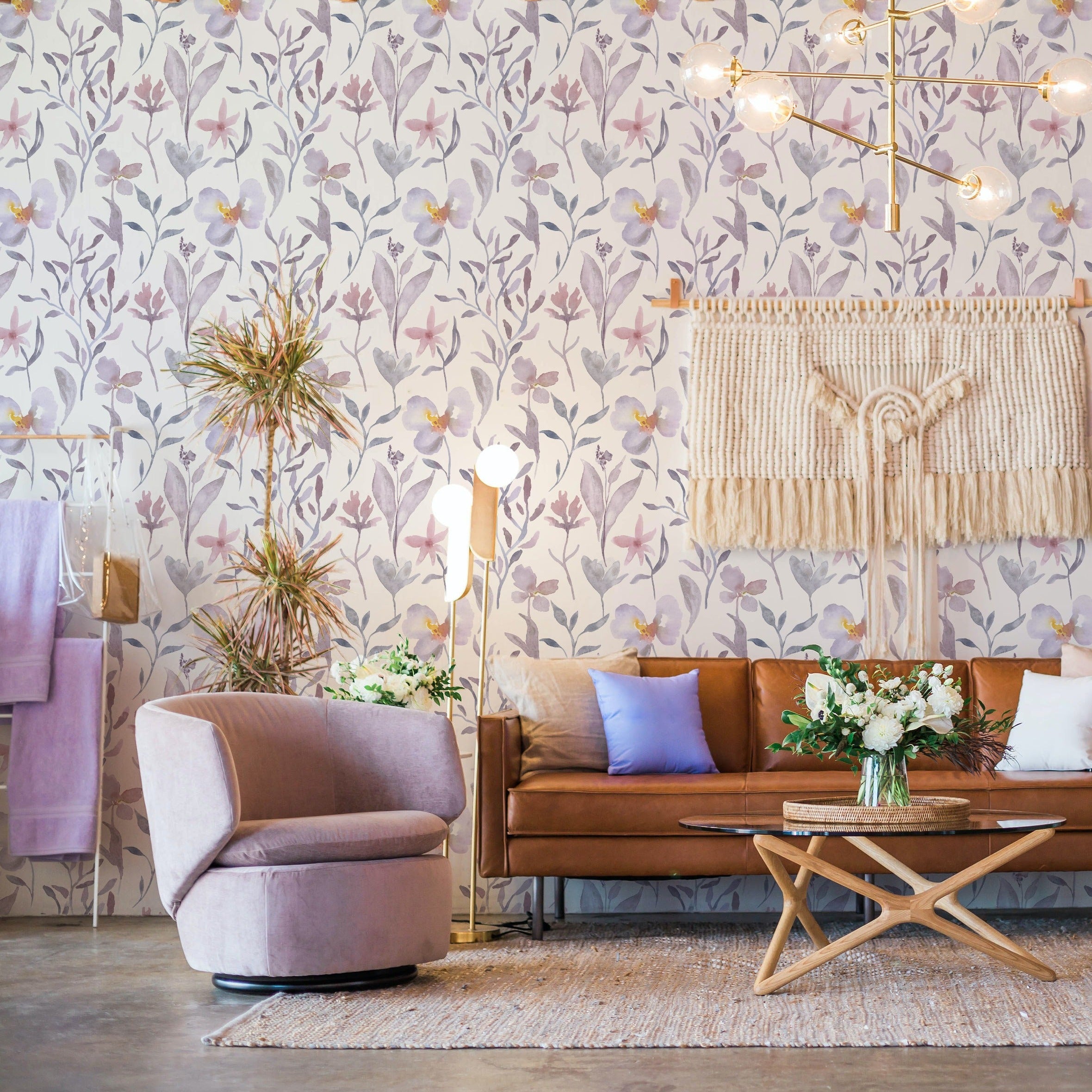 Elegant living room enhanced with Watercolor Garden Wallpaper, showcasing vibrant purple and pink floral designs amidst grey leaves on a white backdrop. The room is furnished with modern decor including a leather sofa, purple chairs, and a wooden coffee table, complemented by soft lighting and a bohemian-style wall hanging, creating a warm and inviting ambiance