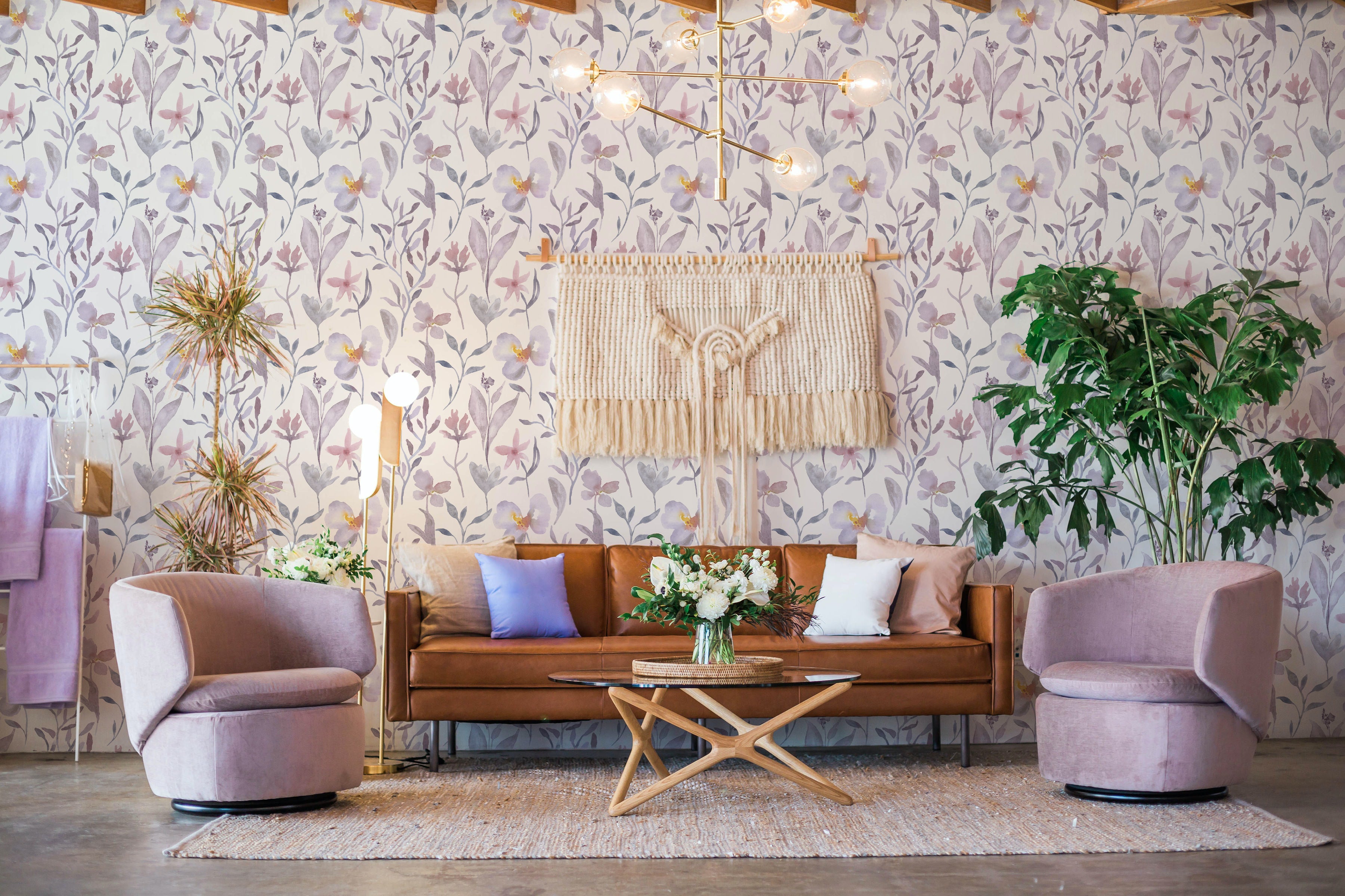 Elegant living room enhanced with Watercolor Garden Wallpaper, showcasing vibrant purple and pink floral designs amidst grey leaves on a white backdrop. The room is furnished with modern decor including a leather sofa, purple chairs, and a wooden coffee table, complemented by soft lighting and a bohemian-style wall hanging, creating a warm and inviting ambiance
