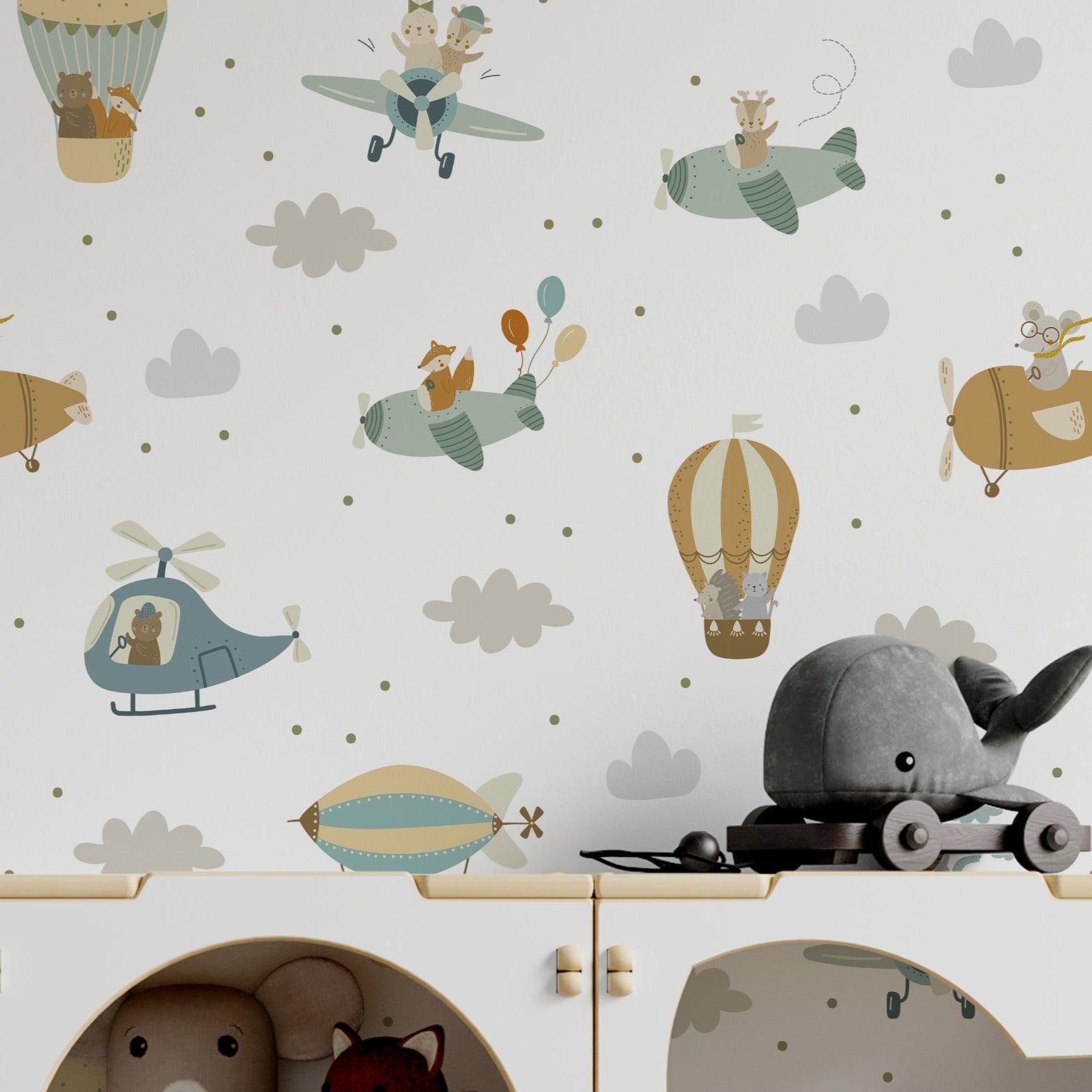Detail of a nursery room wall covered with 'Flying Friends' wallpaper illustrating cartoon animals on airborne adventures. The playful decor includes aviation-themed elements such as hot air balloons, airplanes, and helicopters with animals as pilots amid clouds and stars.