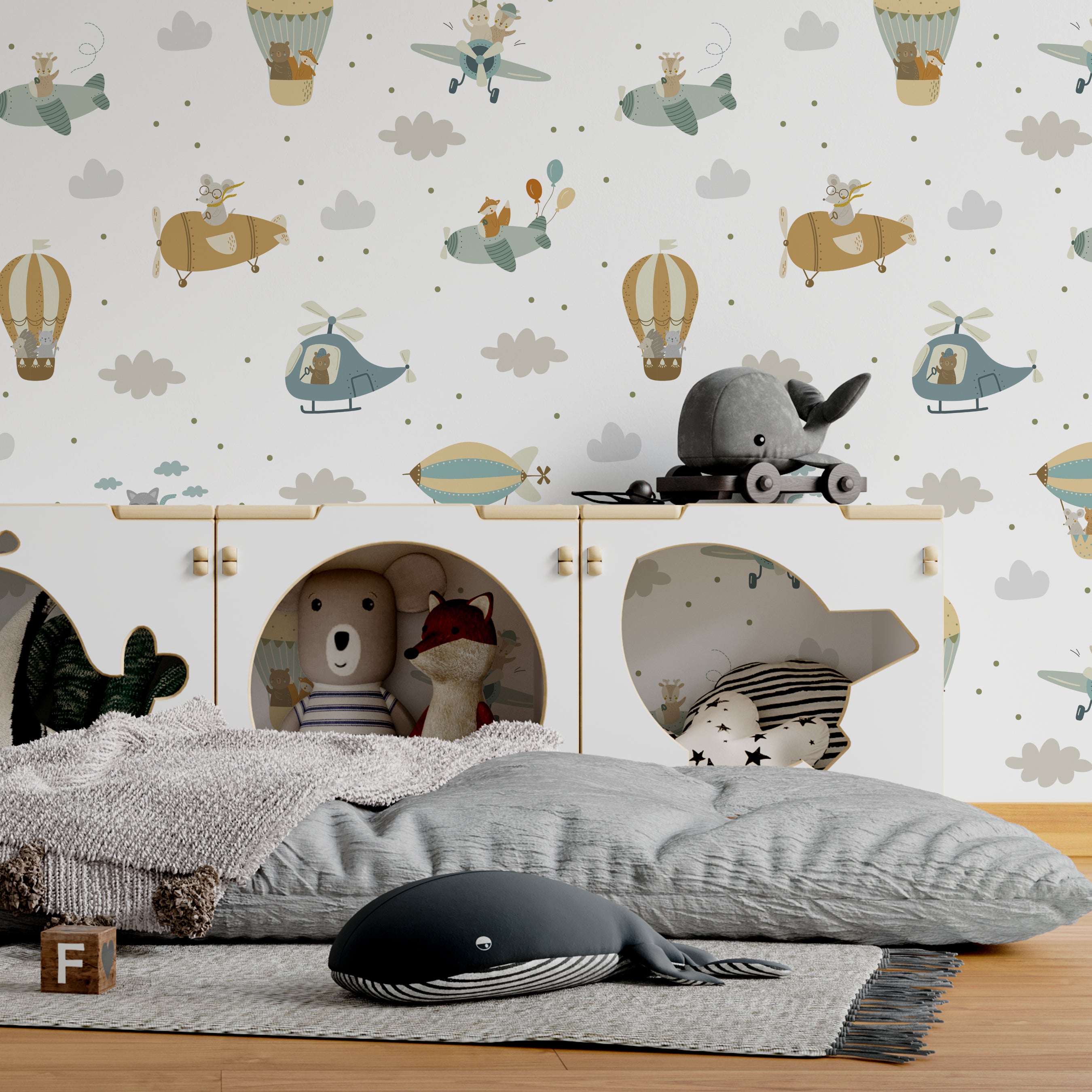 Children's room with 'Flying Friends' wallpaper depicting playful animals piloting air vehicles. A whimsically decorated space features stuffed toys on a two-tiered open cabinet, plush cushions on a bed with grey bedding, and a decorative whale pillow, complementing the adventurous theme of the wallpaper.