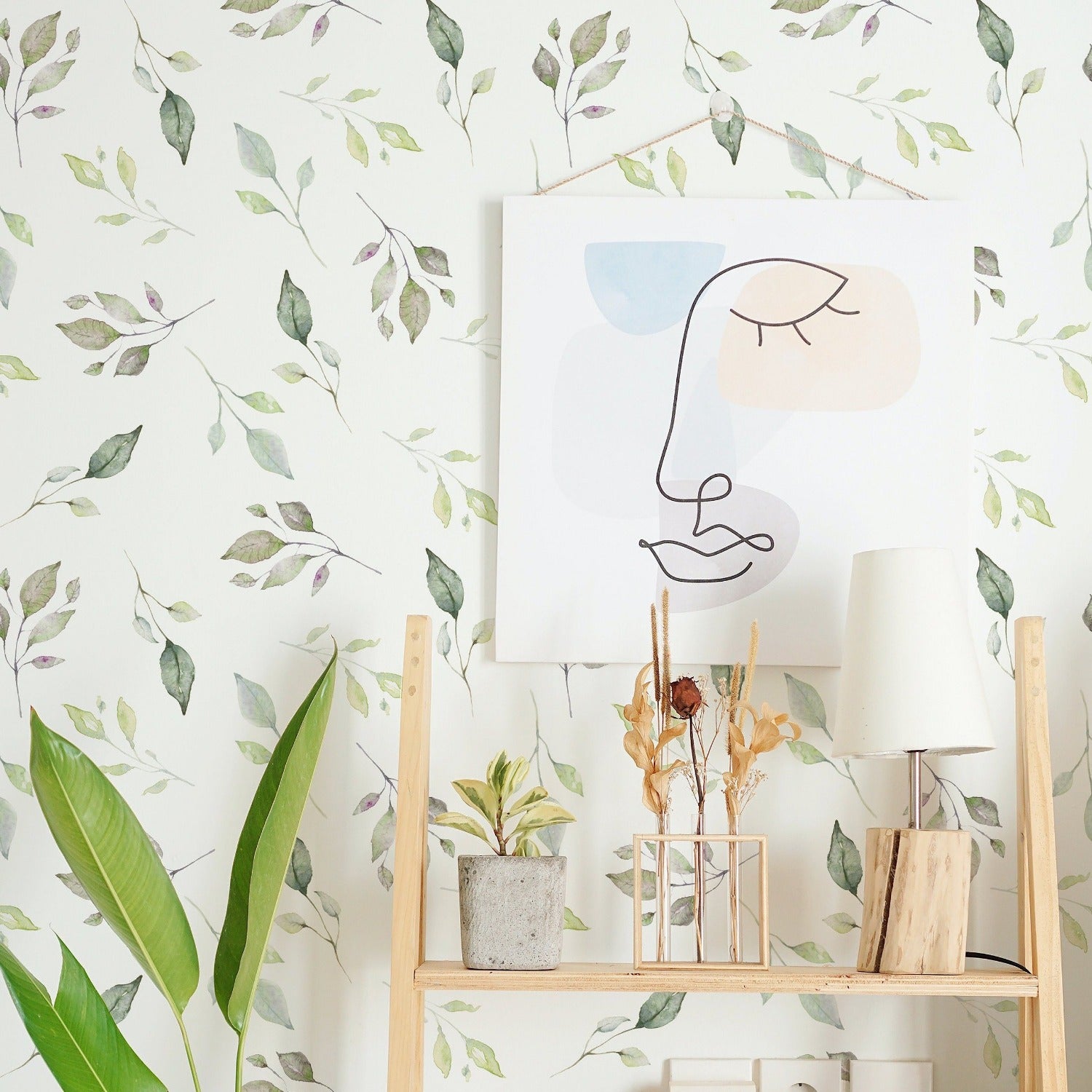 A stylish home workspace with the wall behind featuring 'Fall Floral Wallpaper', a soft watercolor pattern of cascading green leaves. In the foreground, a wooden shelf holds an abstract face drawing, a potted plant, and a table lamp, creating a tranquil and creative atmosphere.