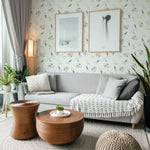 A cozy living room corner with a gray sectional sofa adorned with throw pillows and a tasseled blanket. The wall behind is clad in wallpaper featuring a gentle pattern of watercolor leaves in shades of green on a white background. Two framed abstract art pieces hang on the wall, complementing the serene ambiance.