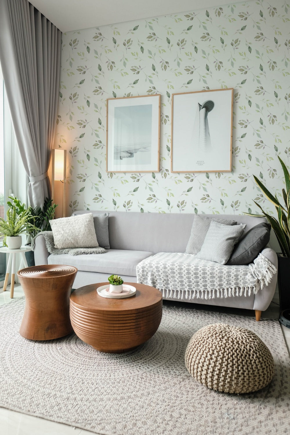 A cozy living room corner with a gray sectional sofa adorned with throw pillows and a tasseled blanket. The wall behind is clad in wallpaper featuring a gentle pattern of watercolor leaves in shades of green on a white background. Two framed abstract art pieces hang on the wall, complementing the serene ambiance.