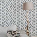 Interior setting featuring a wall adorned with a pale blue floral wallpaper, complemented by a rustic lamp on a wrapped driftwood stand and a patterned cushion on a couch.