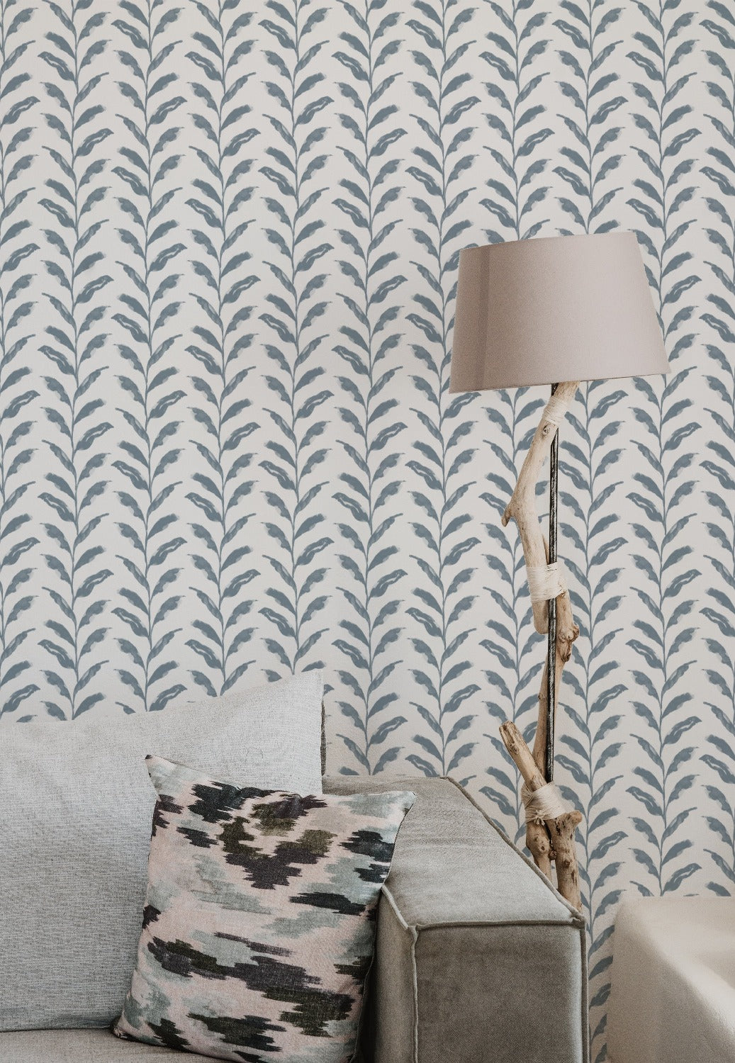 Interior setting featuring a wall adorned with a pale blue floral wallpaper, complemented by a rustic lamp on a wrapped driftwood stand and a patterned cushion on a couch.