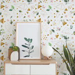 Interior design scene with a wall decorated with a floral and geometric pattern wallpaper, with botanical elements and golden shapes. In front, a wooden shelf holds a framed blank poster, a succulent in a wooden pot, and a spherical textured lamp