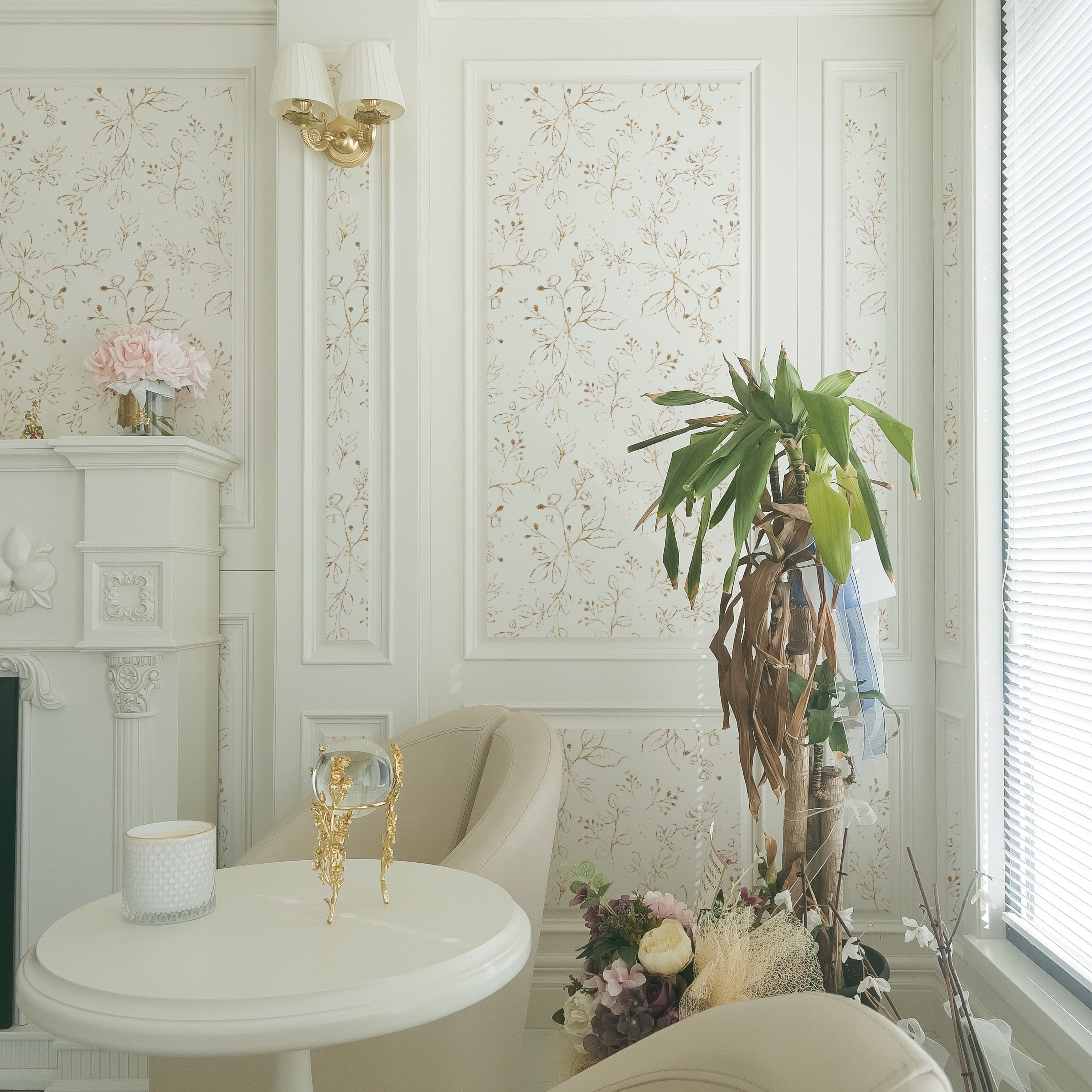 A tranquil corner with a white chair and table against Gold Leaves Wallpaper, creating a luxurious and peaceful reading nook accentuated by warm golden lighting and delicate pink flowers.