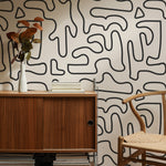A stylish office setup showcasing the Abstract Doodle Wallpaper, with black curving lines and abstract shapes on a neutral beige background. The scene includes a mid-century modern wooden credenza, a contemporary chair, and decorative items like books and a vase with dried leaves, enhancing the wallpaper's artistic appeal.