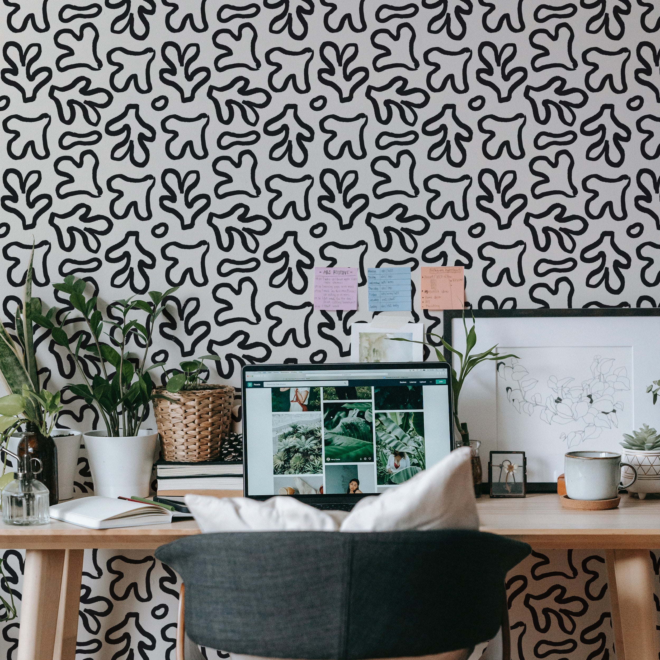 A home office setup showcasing the Bohemian Abstract Wallpaper. The walls are adorned with black organic shapes on a beige backdrop, creating a lively and creative space. The desk area is neatly organized with a computer, books, and green plants, enhancing the room's bohemian vibe.