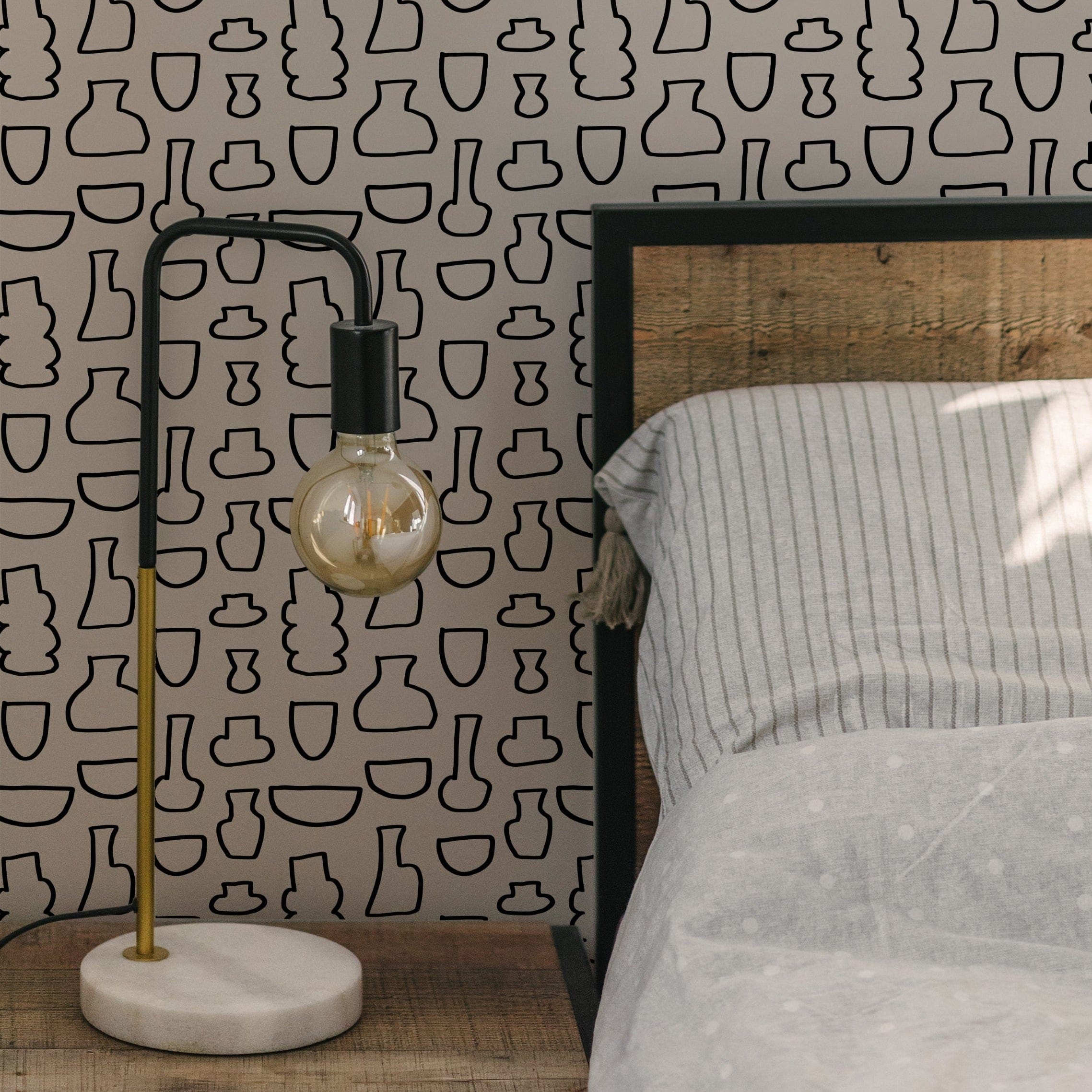 A cozy bedroom setup showcasing the Bohemian Ceramic Wallpaper, adorned with an array of black abstract vase shapes on a light background. The room features a simple bed with striped bedding and a modern lamp, enhancing the bohemian charm of the space.