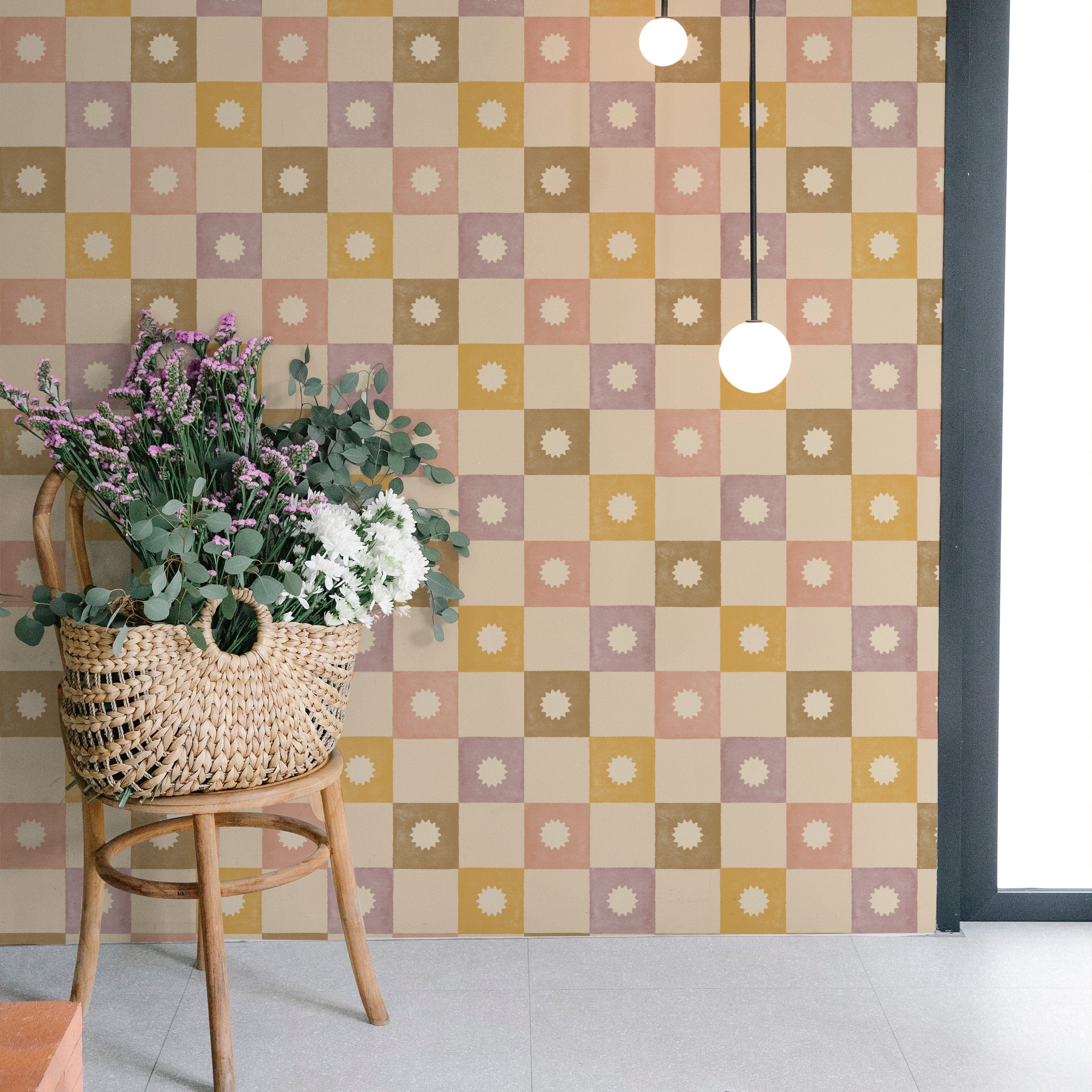 A stylish room corner decorated with Céline Wallpaper, illustrating a pastel checkered design with sunburst motifs. The wallpaper complements a modern setting, enhanced by a rattan chair adorned with fresh flowers, creating a chic and welcoming space