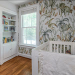 A child's nursery room decorated with the Watercolour Dino Wallpaper, showcasing various dinosaurs and tropical plants in watercolor style. The vibrant scene creates an imaginative and educational backdrop for a white crib and bookshelf