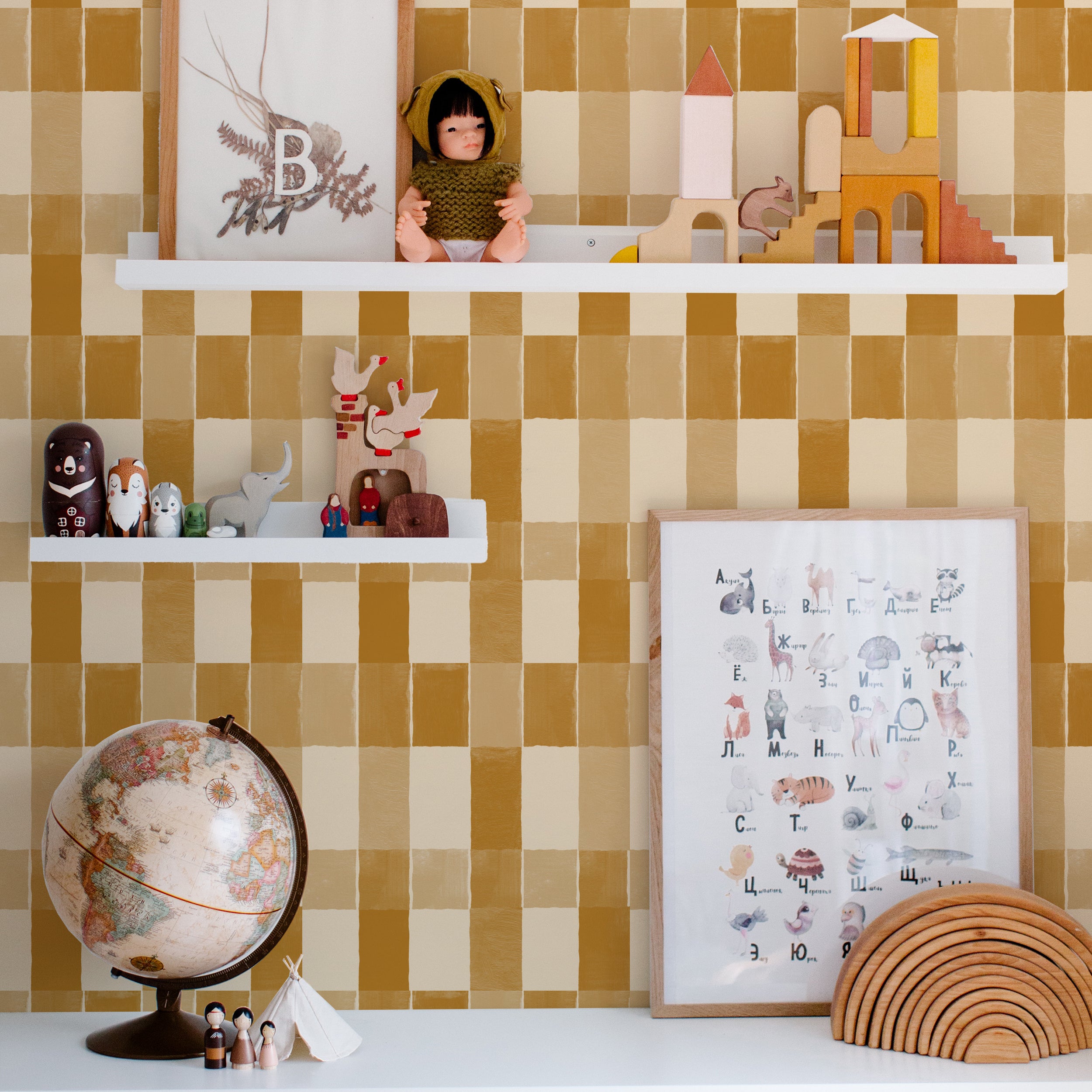 A child's room wall covered with Clara Wallpaper, showcasing a mustard and cream checkered pattern. The wall serves as a backdrop to a white shelf displaying an eclectic mix of toys, decorative objects, and educational posters, creating a vibrant and engaging space