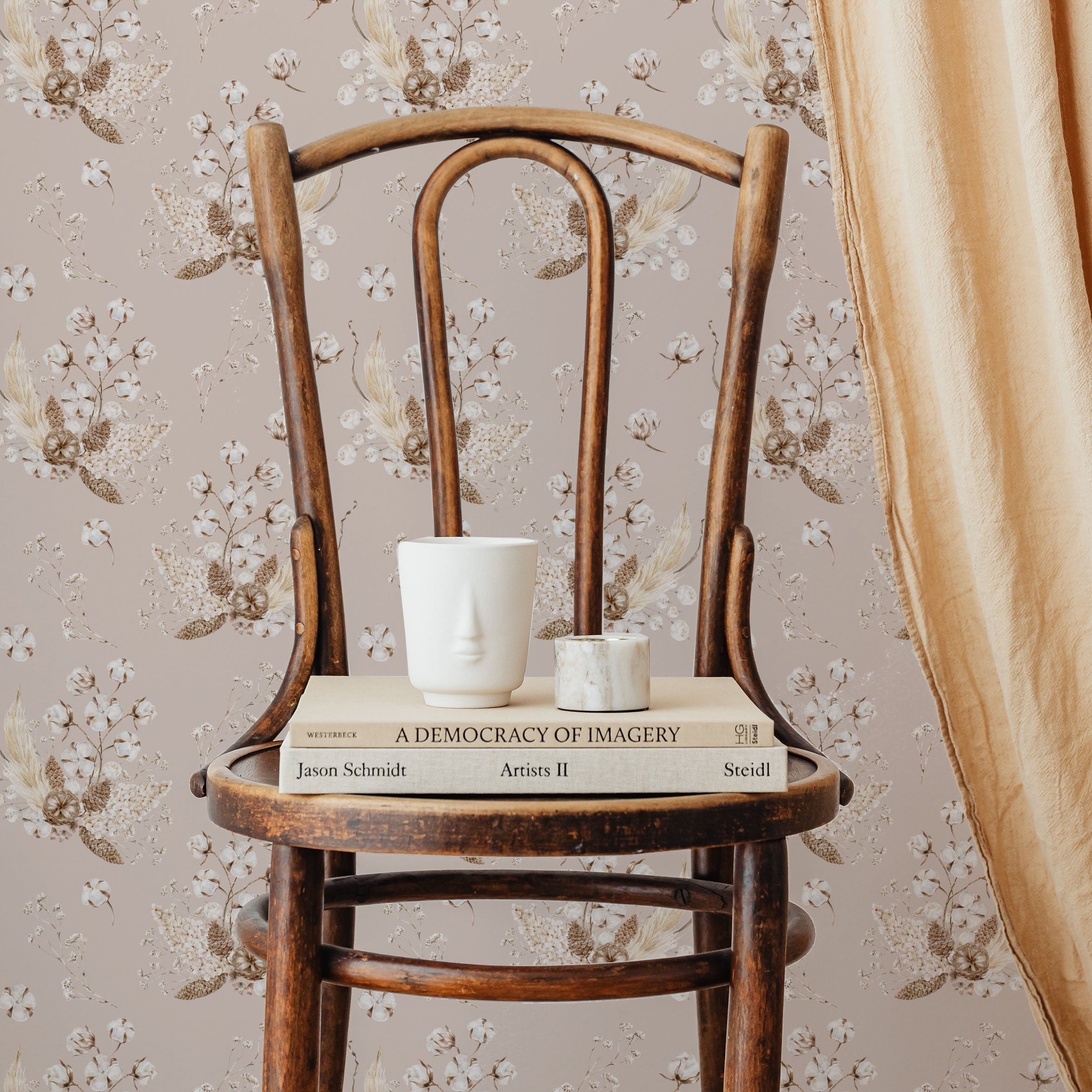 An elegant, vintage wooden chair against a wall covered with 'Boho Bouquet II Wallpaper', featuring clusters of delicate flowers and leaves in soft pinks and golds against a muted pink background. On the chair, a white sculptural face mug and a small marble candle holder sit atop a couple of hardcover books, adding to the room’s bohemian chic aesthetic.