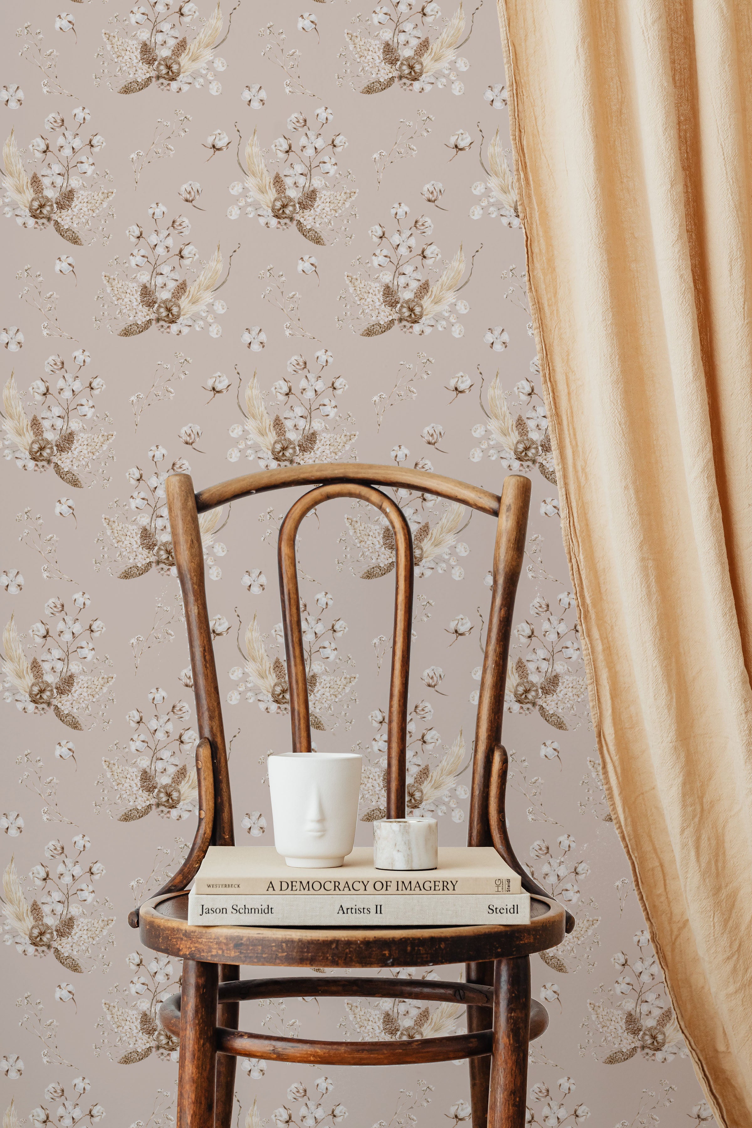 An elegant, vintage wooden chair against a wall covered with 'Boho Bouquet II Wallpaper', featuring clusters of delicate flowers and leaves in soft pinks and golds against a muted pink background. On the chair, a white sculptural face mug and a small marble candle holder sit atop a couple of hardcover books, adding to the room’s bohemian chic aesthetic.