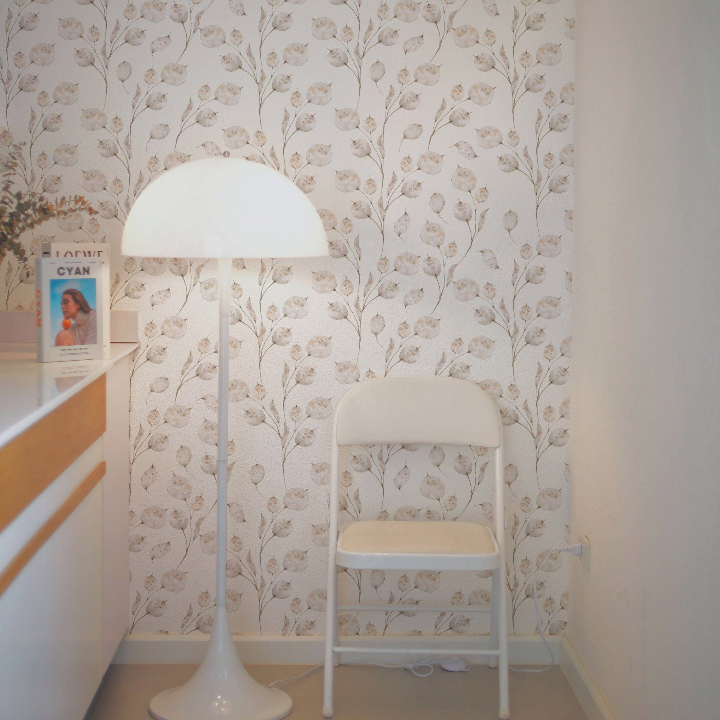 A reading nook with a white chair and lamp, set against a wall decorated with Winter Branches Wallpaper featuring beige botanical designs, creating a serene and stylish atmosphere.