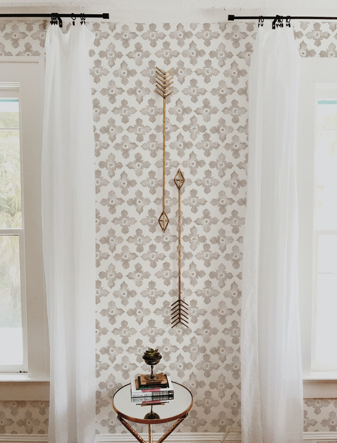 An inviting corner of a room displays the "Moroccan Tile - Beige" wallpaper, which serves as a warm and stylish backdrop for a round side table and a decorative golden arrow wall piece. The gentle beige hues of the wallpaper pattern provide a neutral yet decorative background, illustrating how it can seamlessly integrate into a home's decor.