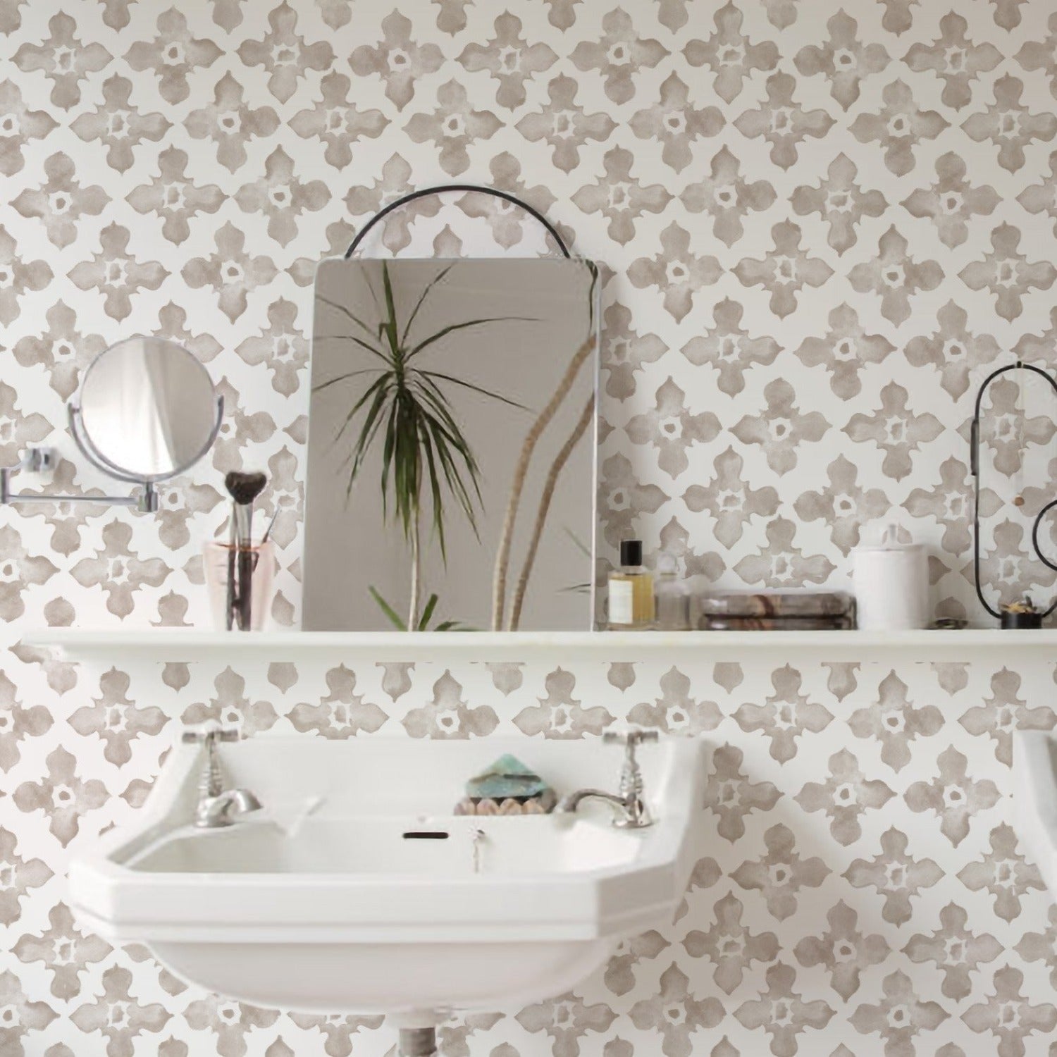 This bathroom interior features the "Moroccan Tile - Beige" wallpaper, with a classic beige and white Moroccan tile pattern that adds an air of sophistication and worldly charm. The wallpaper complements the white pedestal sink and the reflective surface of a rectangular mirror, alongside various bathroom accessories, creating a harmonious and elegant space.