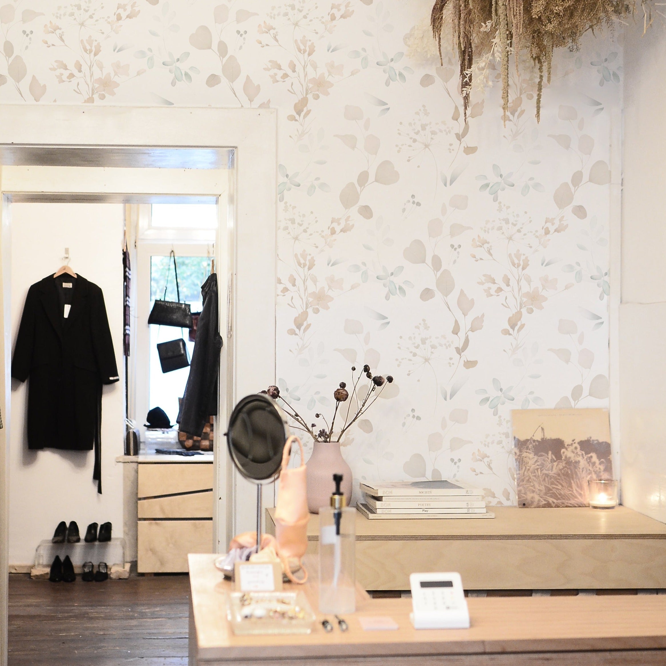 An elegant boutique interior dressed in Watercolour Floral & Herb Wallpaper, complementing the shop's chic decor. The subtle floral pattern serves as a gentle and stylish backdrop to the mirror, clothing rack, and accessories displayed, contributing to a sophisticated shopping atmosphere.
