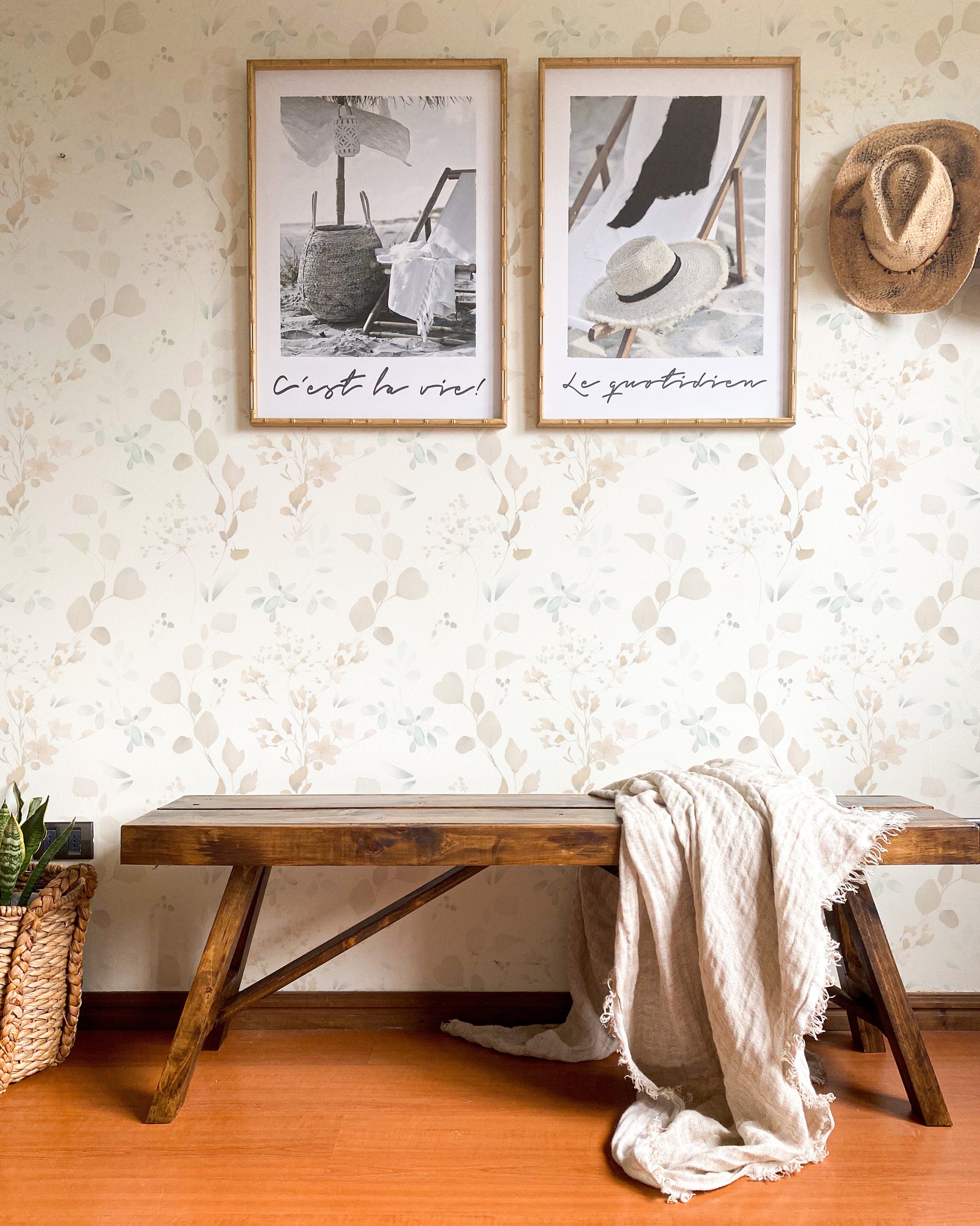 A cozy corner of a room featuring Watercolour Floral & Herb Wallpaper, with pastel botanical patterns creating a tranquil backdrop. The scene includes a rustic wooden bench with a draped cream throw blanket, two framed beach-themed artworks, and a straw hat, suggesting a relaxed, coastal-inspired interior.