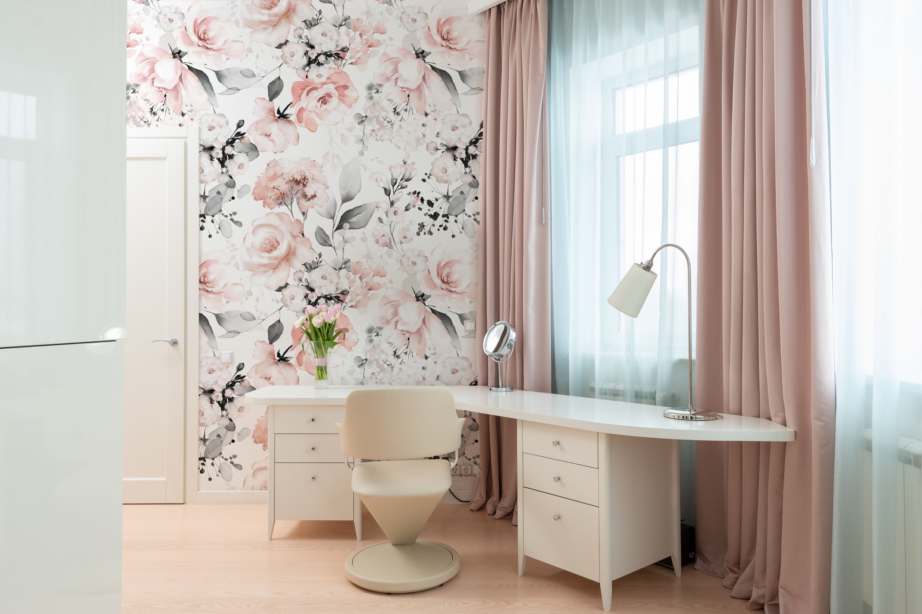 A bright and inviting beauty corner with 'Floral and Herbs Wallpaper - Pink + Black - 50"' adorning the wall. The wallpaper features large, soft pink florals and muted black herbs on a clean white background, complementing the pastel pink curtains and the modern white vanity setup with a stylish cream chair.