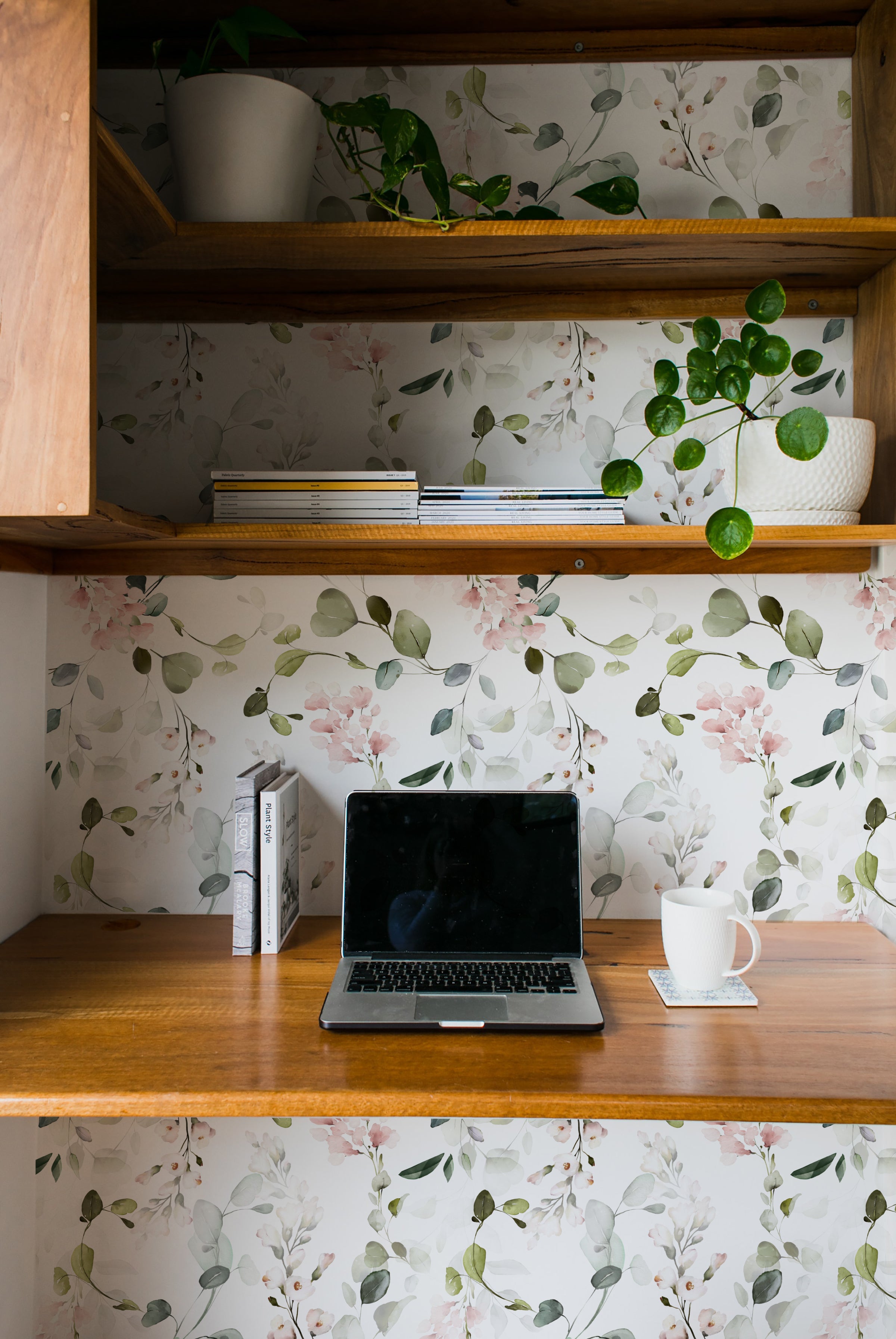 A cozy home office setting, the Pink Floral & Herbs Wallpaper creating a tranquil backdrop behind a wooden desk. The space is accented with a potted plant, stacks of books, and a laptop, crafting a harmonious blend of nature-inspired design and productivity.