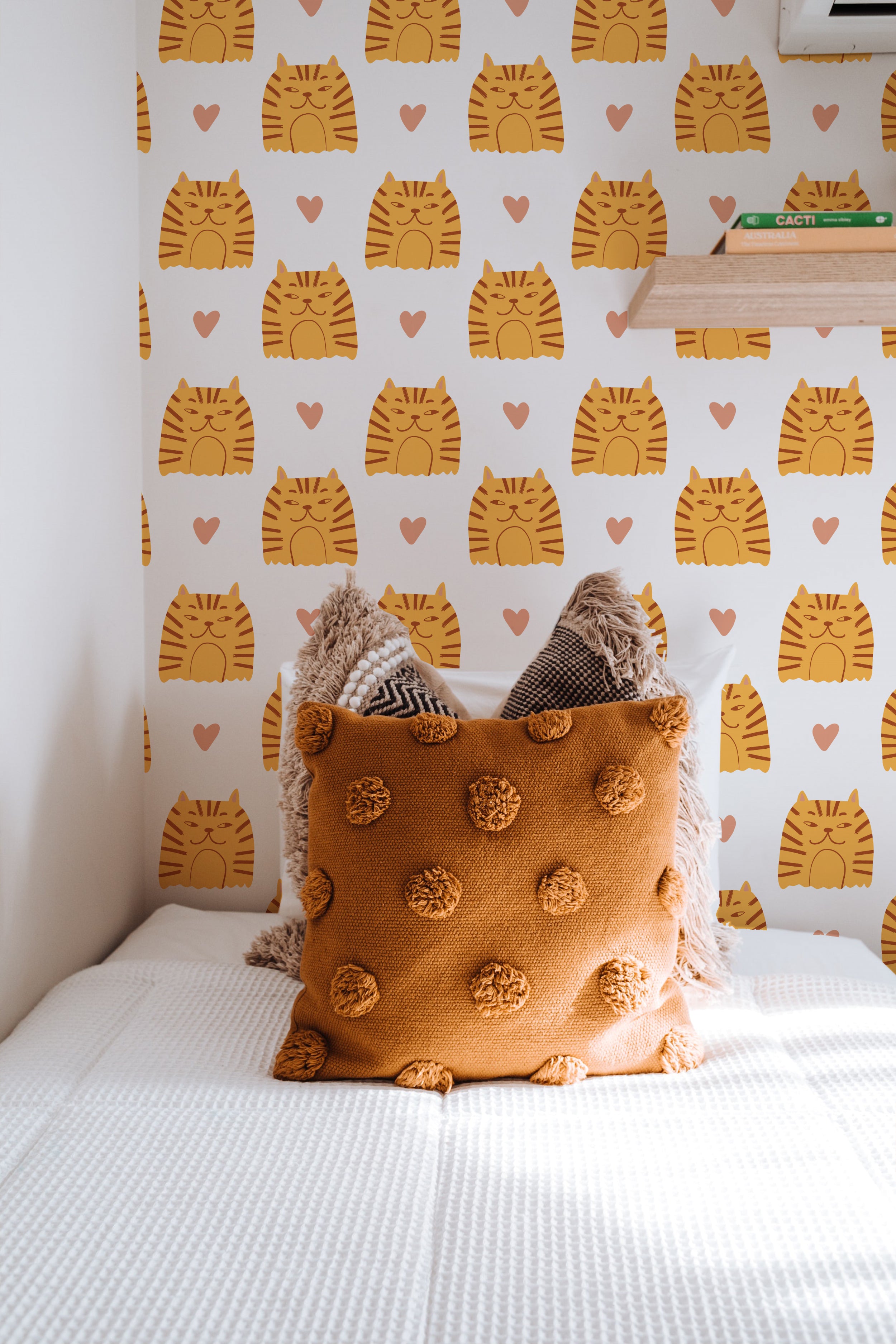 A cozy bedroom setting featuring the 'Cat Wallpaper 9' adorned with orange striped cats and pink hearts. The room includes a stylish mustard textured pillow and matching blankets, enhancing the warmth and inviting nature of the space