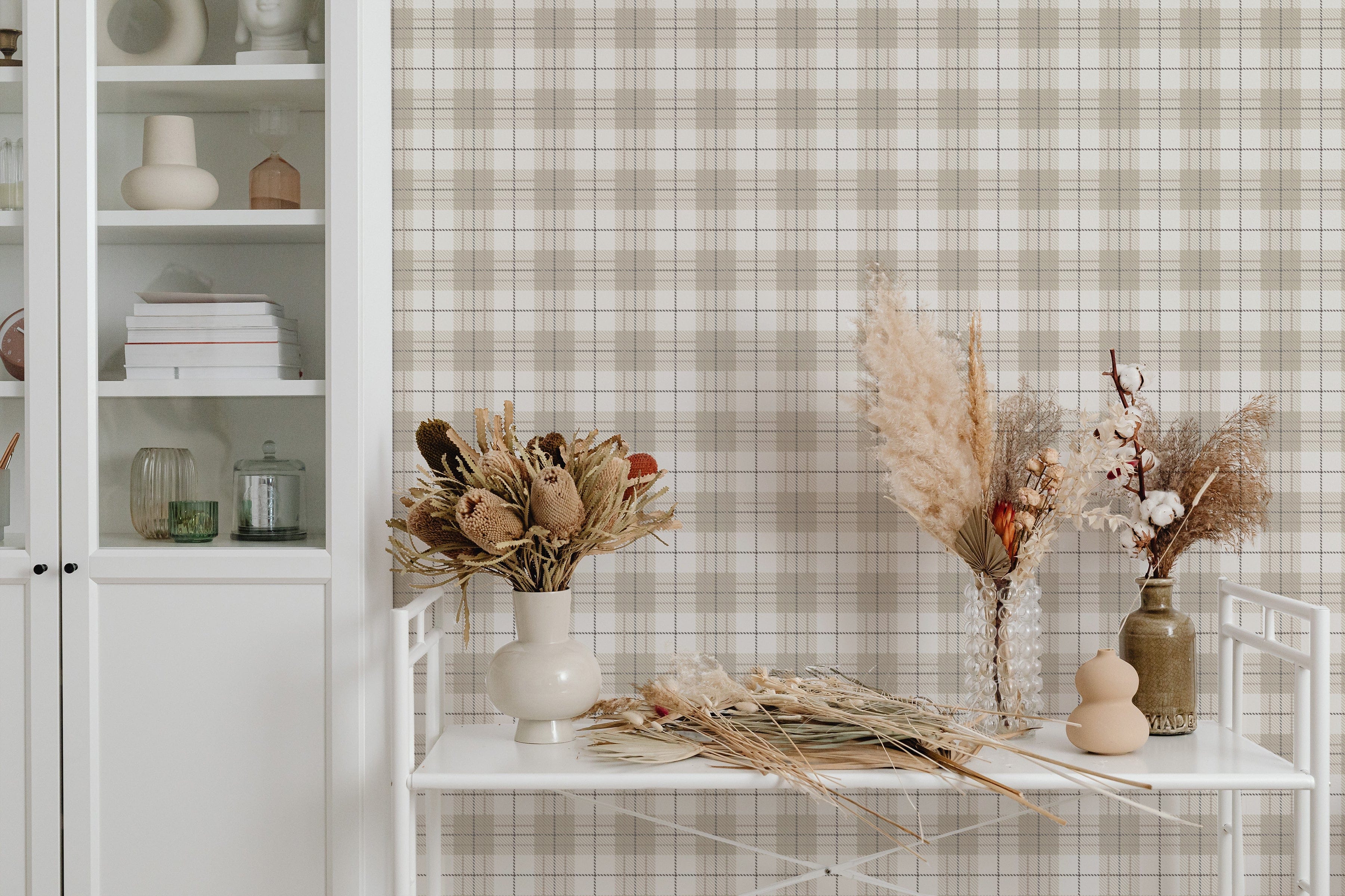 A bright and airy corner of a room featuring Winter Plaid Wallpaper with a large bookshelf and a side table decorated with dried flowers. The wallpaper's subtle plaid pattern in beige and white offers a cozy backdrop that complements the natural textures and soft color palette of the decor.