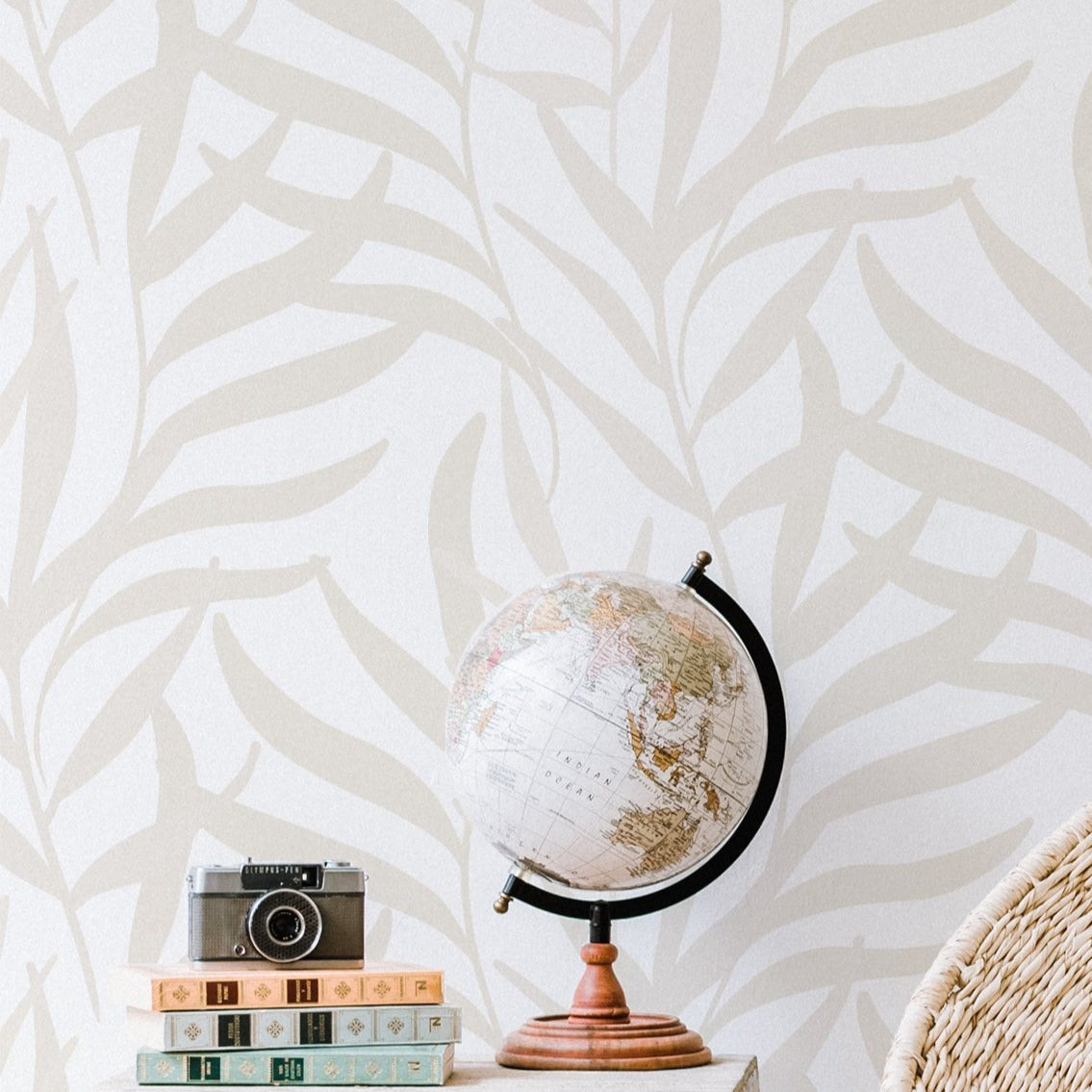 A cozy corner of a room featuring a globe on a wooden stand, placed atop a stack of decorative books beside a vintage camera. The backdrop is a neutral-toned wall adorned with a subtle, leafy 'Earthy Wallpaper' pattern, creating a tranquil and cultured ambiance.