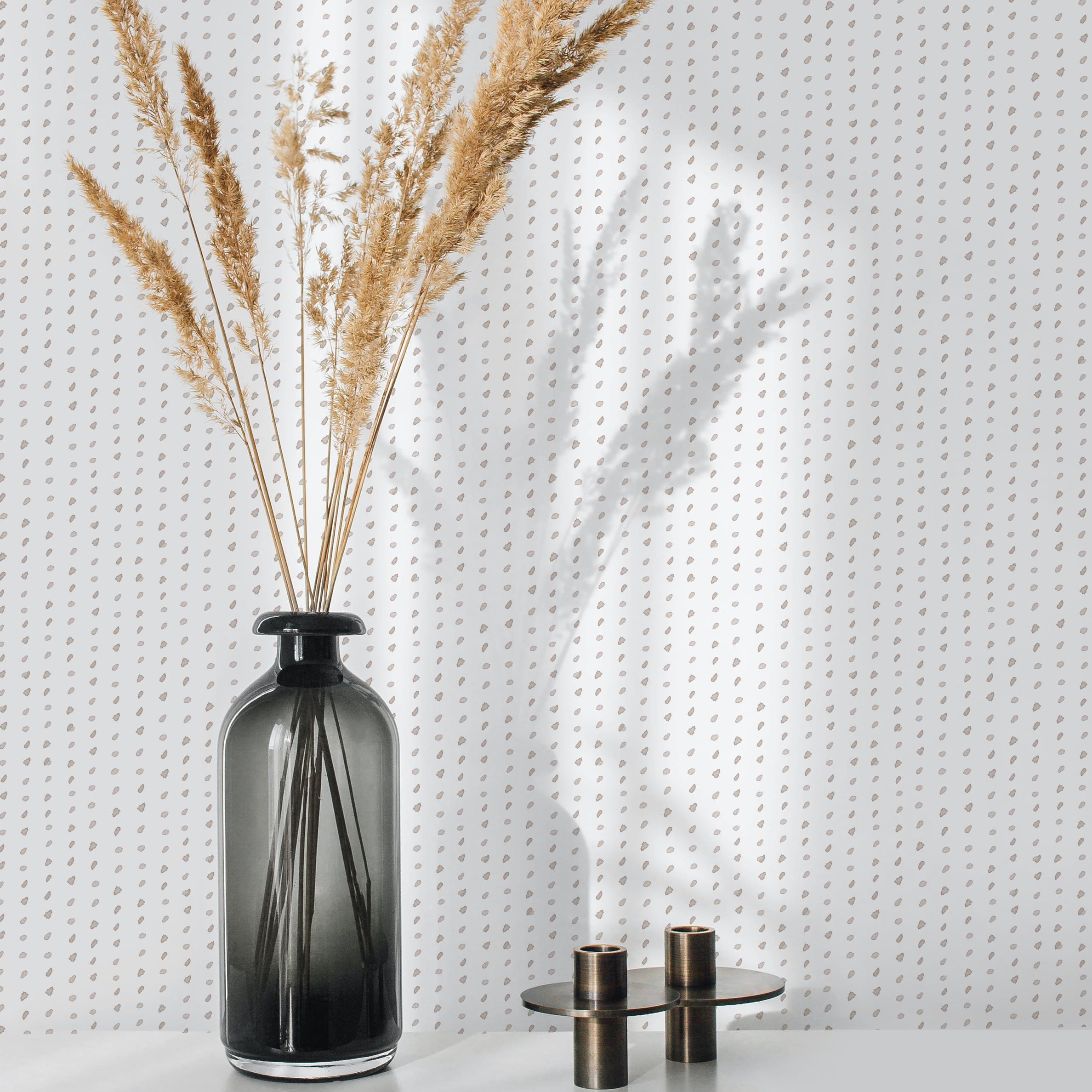 A square image showing a room vignette with a section of wall covered in a minimal watercolor wallpaper. The design comprises tiny, dark teardrop shapes set against a clean white backdrop. A sleek black vase with tall, dried beige grasses and a small brass candle holder set add a sophisticated touch to the scene.