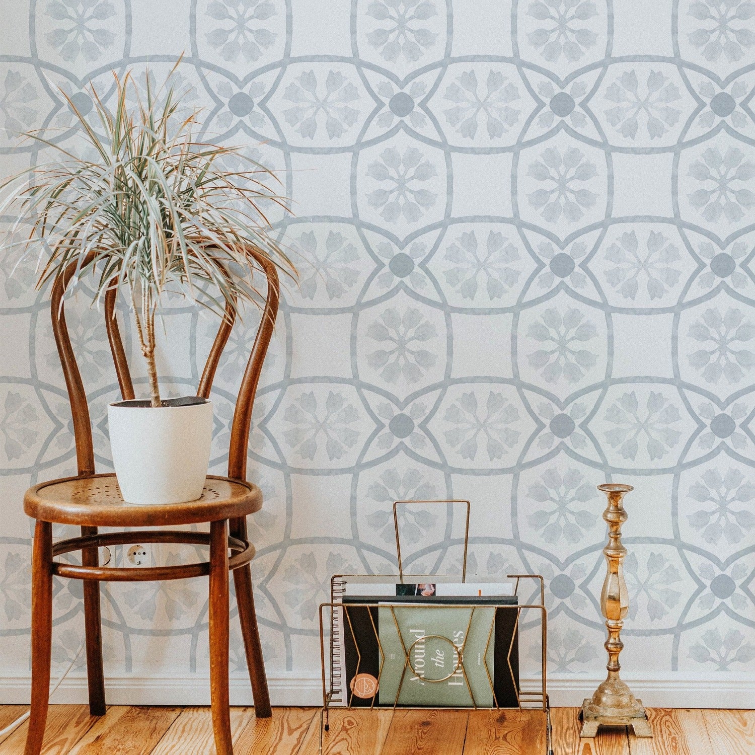 A cozy corner showcasing the 'Distressed Moroccan Tile - Pale Blue' wallpaper in a real-life setting. The subtle pale blue and grey tones create a tranquil ambiance, complemented by a vintage wooden chair, a potted plant, and antique decorative items that enhance the Moroccan-inspired aesthetic