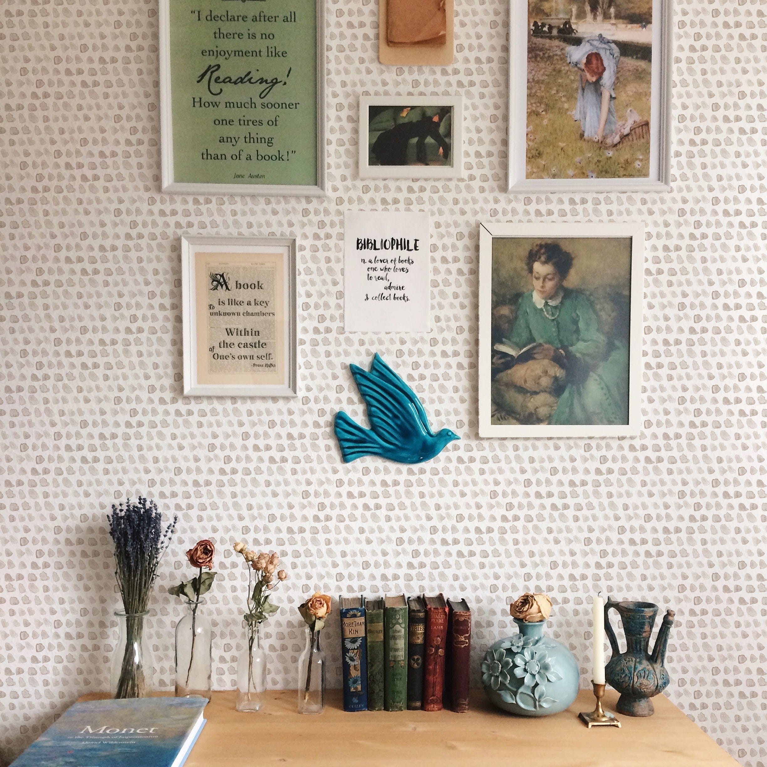 A cozy room corner decorated with Hand Painted Dots Wallpaper - Beige, adorned with various wall hangings and a collection of vintage books and decorative items on a wooden shelf, showcasing the wallpaper's compatibility with warm, inviting decor.