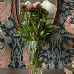 Peacock Damask Wallpaper in a sophisticated interior, featuring a dark floral and peacock design with elegant pink and green accents, a vintage mirror, and a glass vase with fresh peonies on a wooden table.