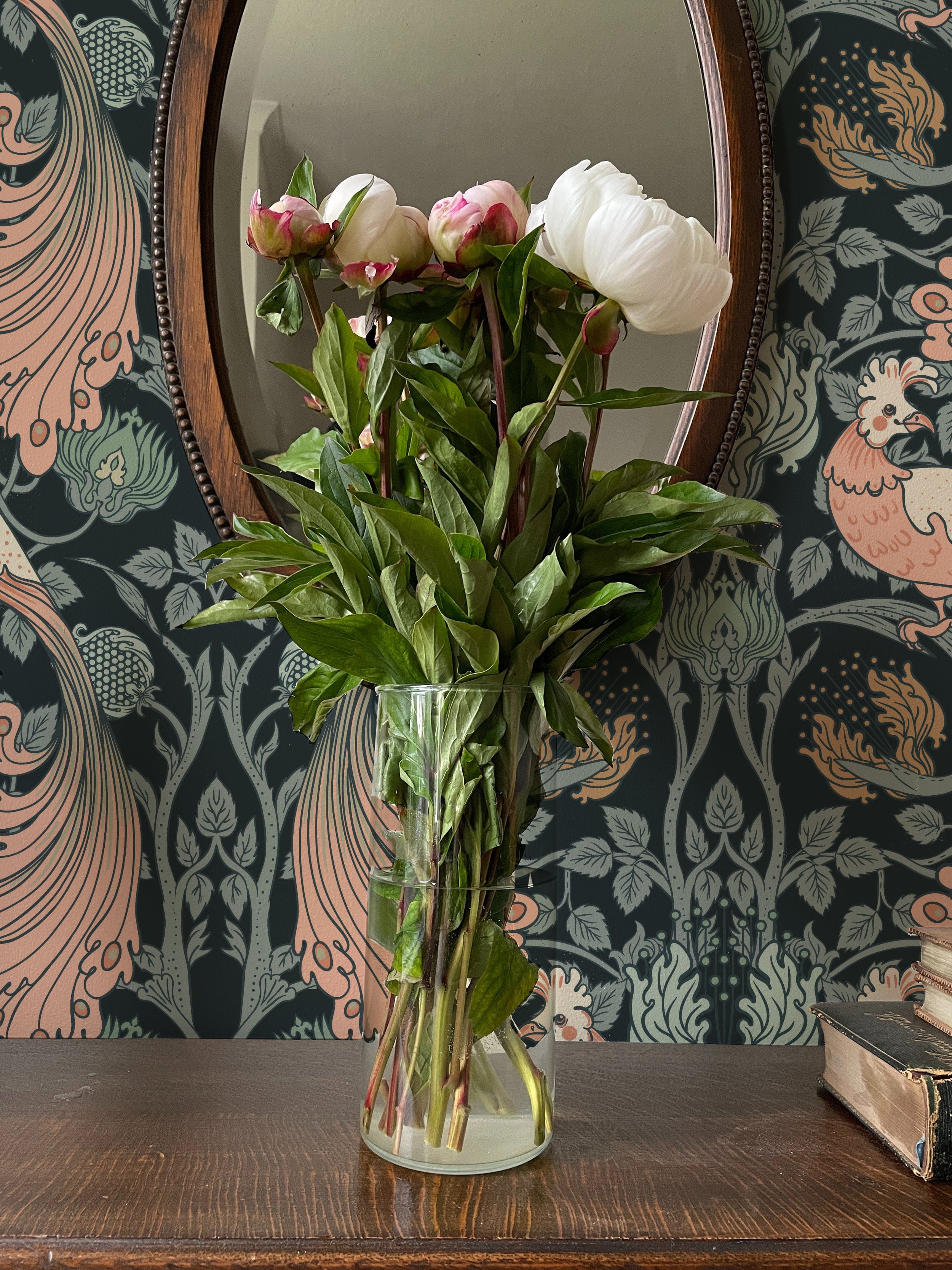 Peacock Damask Wallpaper in a sophisticated interior, featuring a dark floral and peacock design with elegant pink and green accents, a vintage mirror, and a glass vase with fresh peonies on a wooden table.