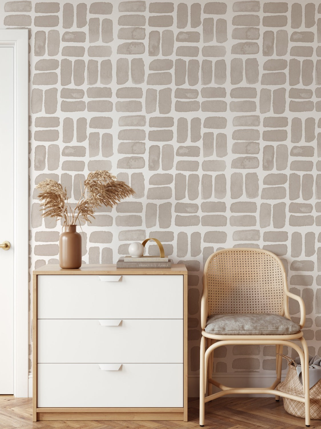 A cozy corner of a room featuring the 'Sea Breeze Wallpaper', with a hand-painted style beige brick pattern giving a soft, textured look, accompanied by a modern white dresser and a rattan chair with a cushion.