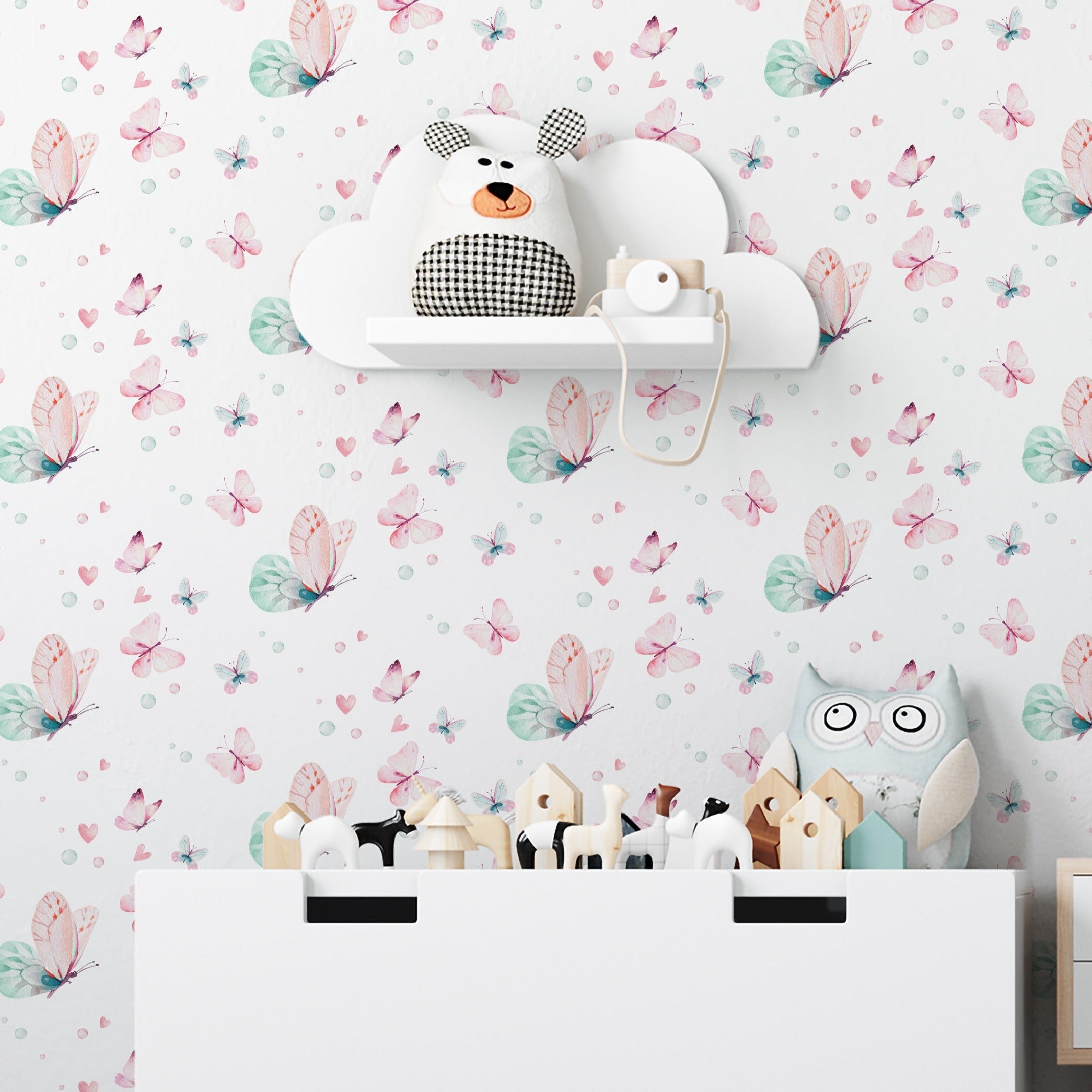 A modern nursery room showcasing the 'Fairy and Flowers III' wallpaper adorned with pink and teal butterflies and small floral accents. The room features contemporary white furniture and children’s toys, complementing the delicate and whimsical nature of the wallpaper.