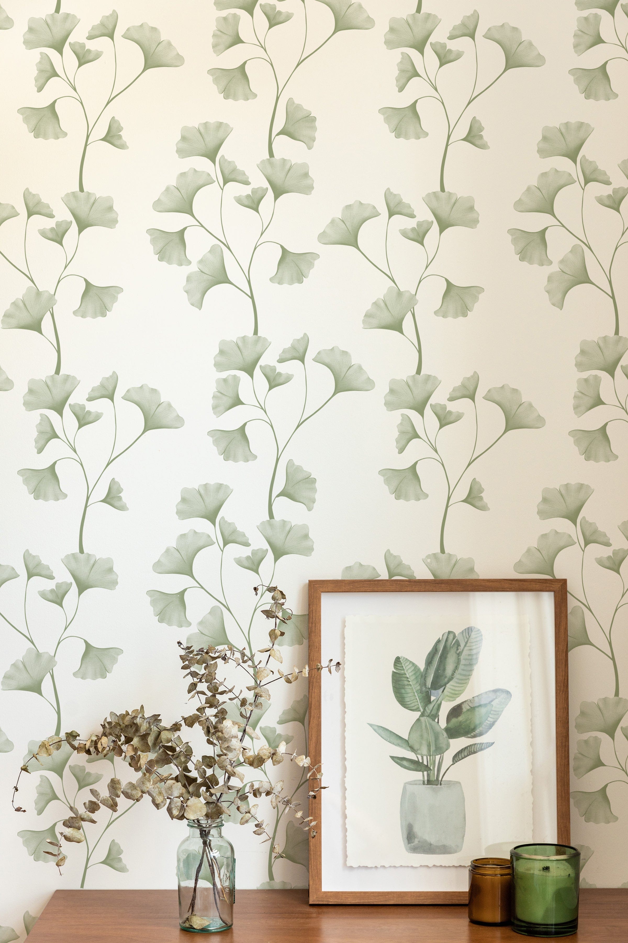 A home decor scene featuring the Botanical Vines Wallpaper, which displays elegant, stylized vine patterns with soft green leaves on a cream background. The setting includes a wooden frame with a botanical art print and decorative green glass vases enhancing the natural theme.